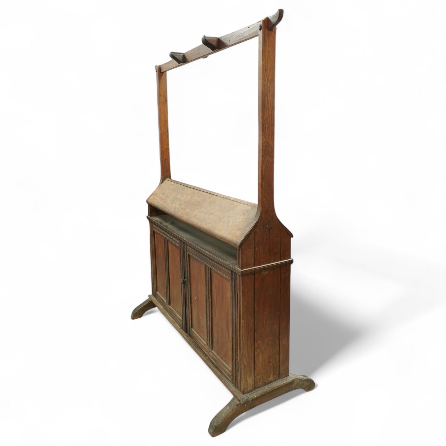 Early 20th century oak saddle and tack stand, the sloped top section for saddle storage raised on rectangular panelled base with cupboard doors enclosing shelved interior, with integral upright frame and horizontal rail fitted with three curved tack hook