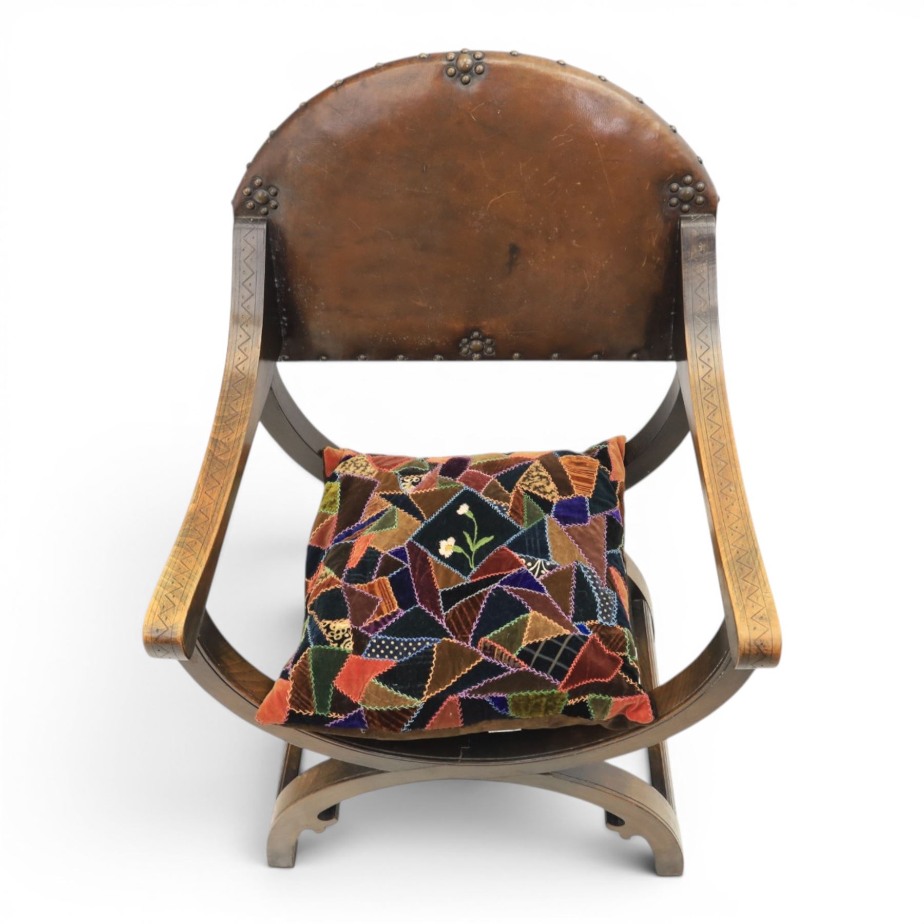 Early 20th century mahogany Savonarola chair, arched leather back with brass studwork, curved open arms with incised geometric decoration, loose patchwork velvet cushion seat, X-frame supports united by circular side rails, on scrolled feet