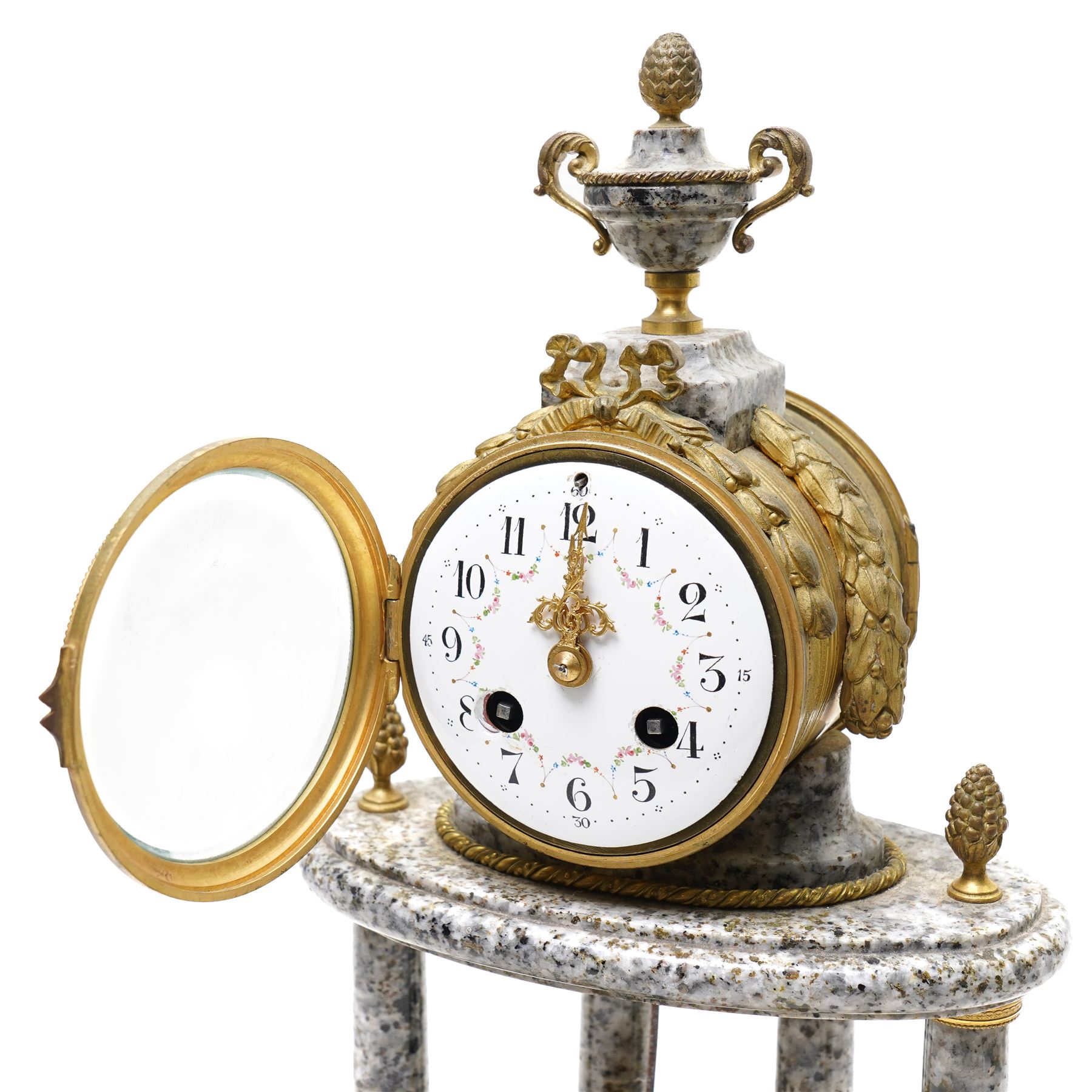 French- early 20th century portico clock garniture, 8-day drum movement decorated with laurel leaves to the glazed bezel and surmounted by a decorative urn with scroll handles, raised by four matching columns on a granite effect oval base with toupee feet, convex enamel dial with gilt pierced hands, Arabic numerals, minute markers and a garland of flowers, twin train Parisian rack striking movement, striking the hours and half hours on a bell. With a sunburst pendulum and key and a matching pair of two-light candelabra.
