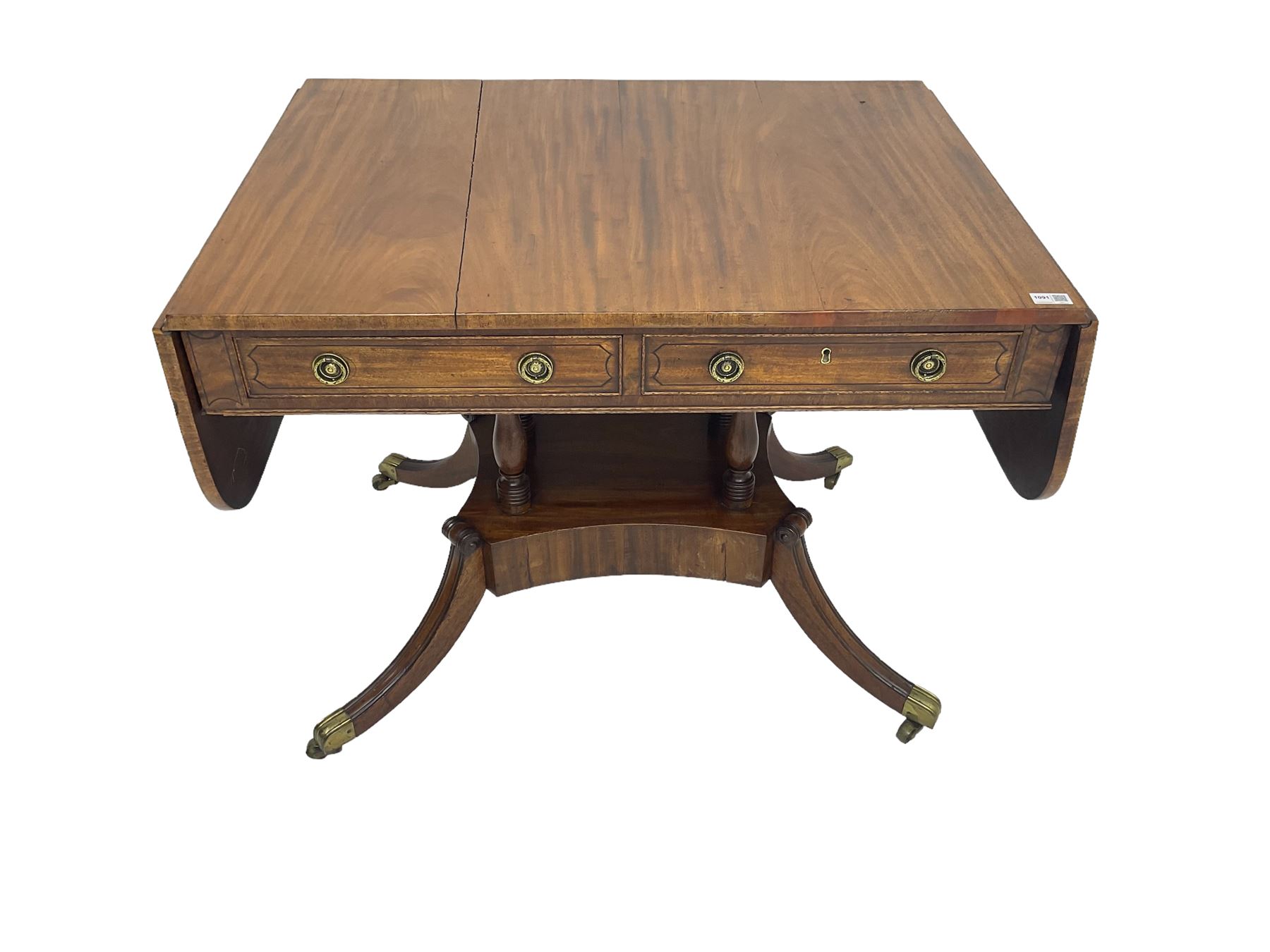 Regency period mahogany drop-leaf sofa table, rectangular top fitted with two cockbeaded drawers with ebony stringing, raised on turned columns united by quadrupod base, the sabre supports terminating in brass cups and castors