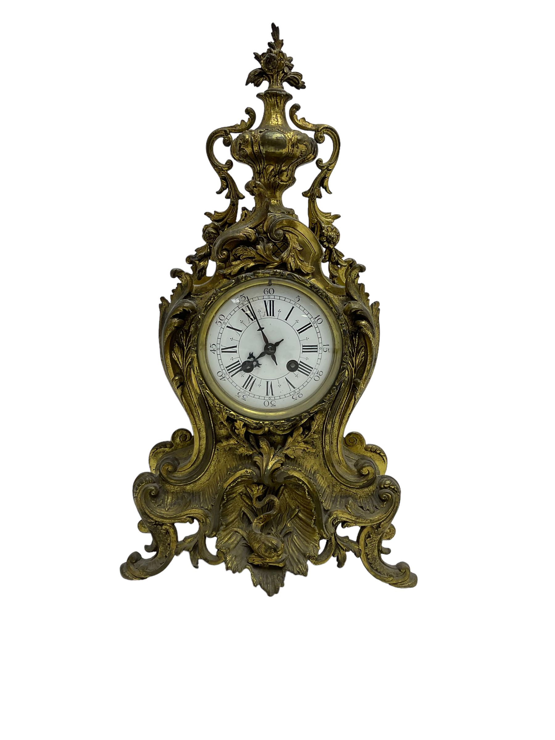 French - late 19th century 8-day gilt brass mantle clock surmounted by an urn, with a waisted case and baroque decoration raised on four splayed feet, with a glazed bezel, enamel dial with Roman numerals, five minute Arabic's and gothic steel hands, count wheel striking movement, striking the hours and half-hours on a bell. With pendulum.