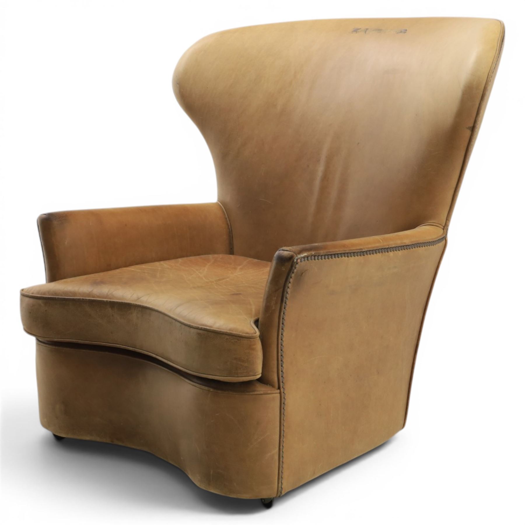 20th century tan leather armchair, the curved wing back above low square arms, shaped loose seat cushion with piped edge, close-nailed side detail, raised on concealed castors