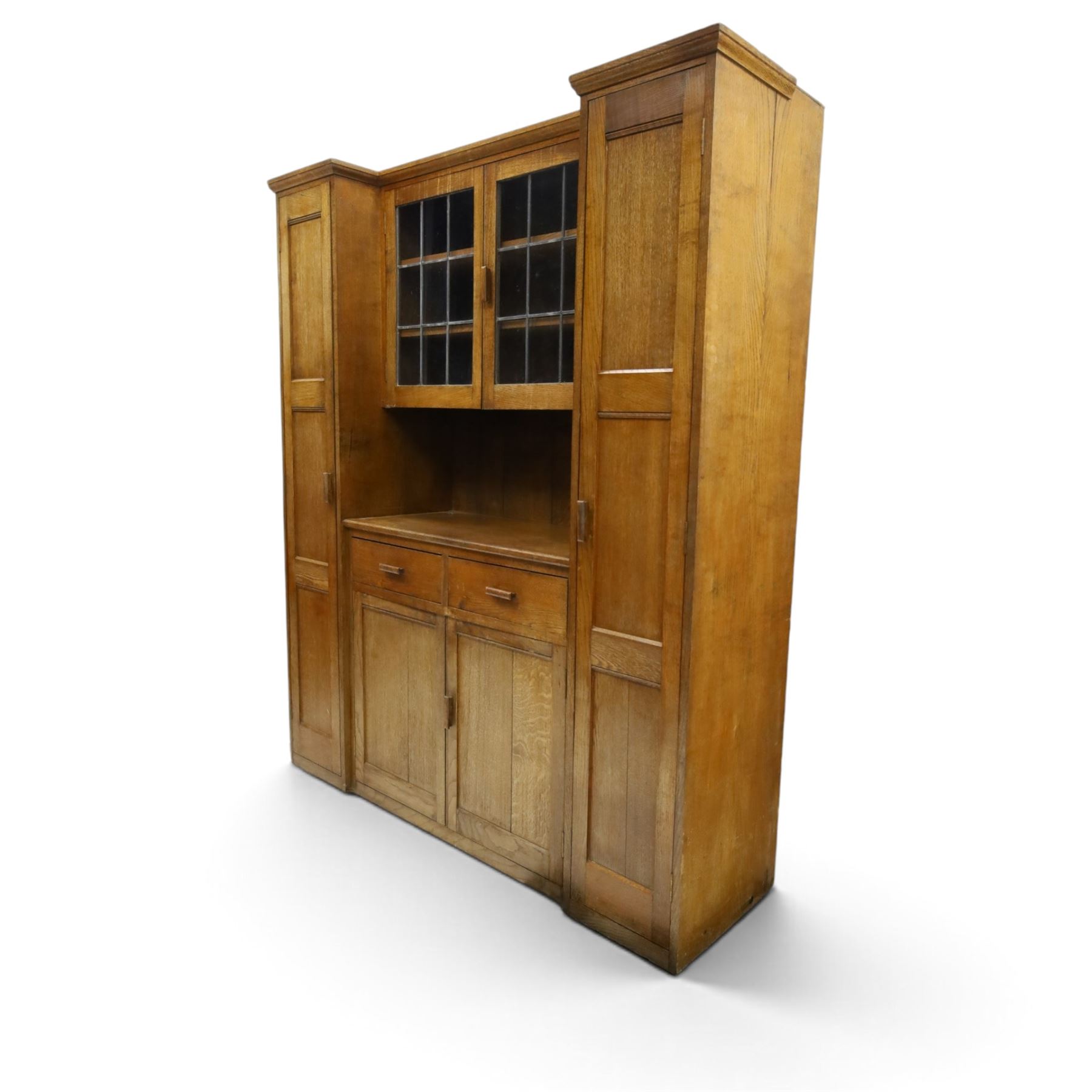 Arts & Crafts oak housekeeper’s cupboard, stepped moulded cornice over twin leaded glazed doors enclosing shelved interior, flanked by narrow panelled cupboards, open recess below over two drawers and twin panelled cupboard doors, with rectangular handles