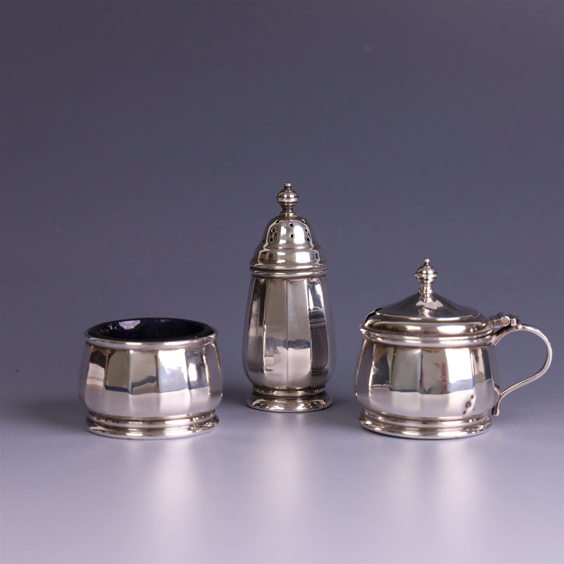 Mid 20th century three piece silver cruet set, comprising pepper shaker, open salt and mustard pot with cover, each of circular faceted form, hallmarked Walker & Hall, Birmingham 1958, the mustard pot and salt with blue glass liners