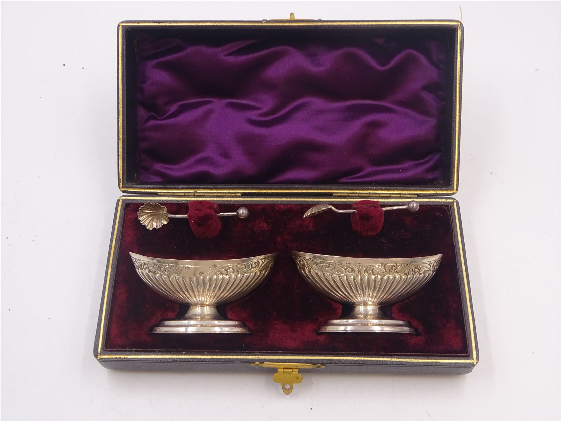 Pair of Victorian silver open salts, of part fluted navette form, with engraved foliate decoration, upon oval stepped foot, hallmarked W H Lyde, Birmingham 1896, H3.5cm, together with a pair of silver salt spoons, with leaf shaped bowls and ball finials, hallmarked Joseph Gloster, Birmingham 1897, together with a pair of Victorian silver open salts, of circular form, with shaped rim and embossed scroll and lattice decoration to the gilded bowl, together with pair of matching silver salt spoons, hallmarked Albert Sydenham, Birmingham 1899, both sets within tooled leather, velvet and silk lined fitted cases