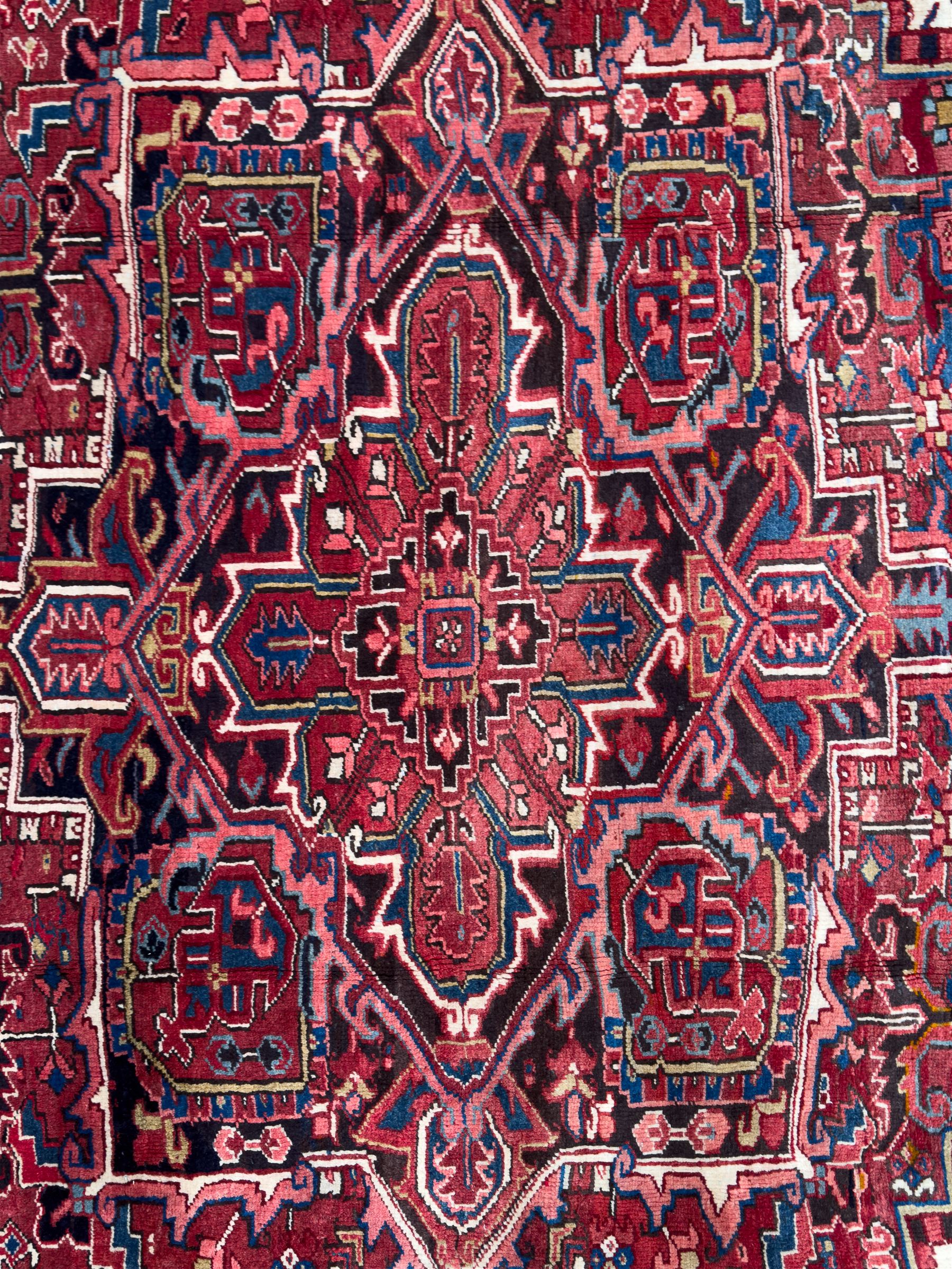 Northwestern Persian Heriz hand-knotted carpet, red madder field with a bold stepped medallion in indigo, ivory, and terracotta, flanked by angular floral motifs and stylized leaves, the spandrels set within a dark indigo border of rosettes and angular vine motifs between narrow guard stripes, 340cm x 245cm 