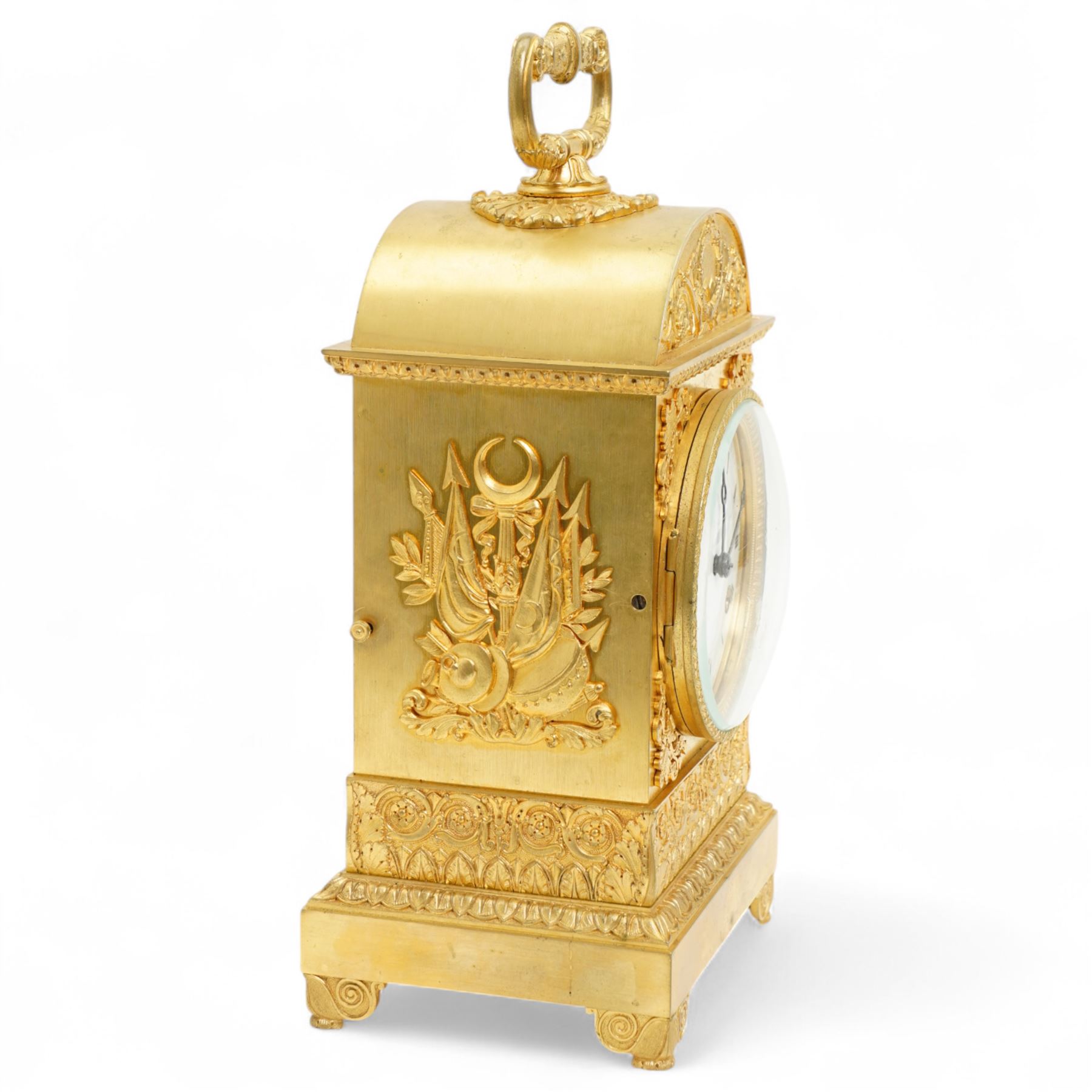 Fine Swiss ormolu 8-day early 19th century Pendule d'Officer style clock c1820,  circular double train ting-tang movement striking the quarters on a graduated pair of bells and sounding the hour on one, going train with a vertical platform lever escapement and over sprung balance, rack striking, with visible hour and quarter strike work to the backplate, movement unsigned, convex enamel dial with Roman numerals and steel moon hands behind a glazed gilt bezel, case with a scroll cast handle to the arched top with applied rococo decoration to the tympanum, both sides of the case adorned with a military trophy of arms motif and deep frieze with foliate arcading beneath, on a rectangular plinth with decorative scroll engraved bracket feet. With Key.   