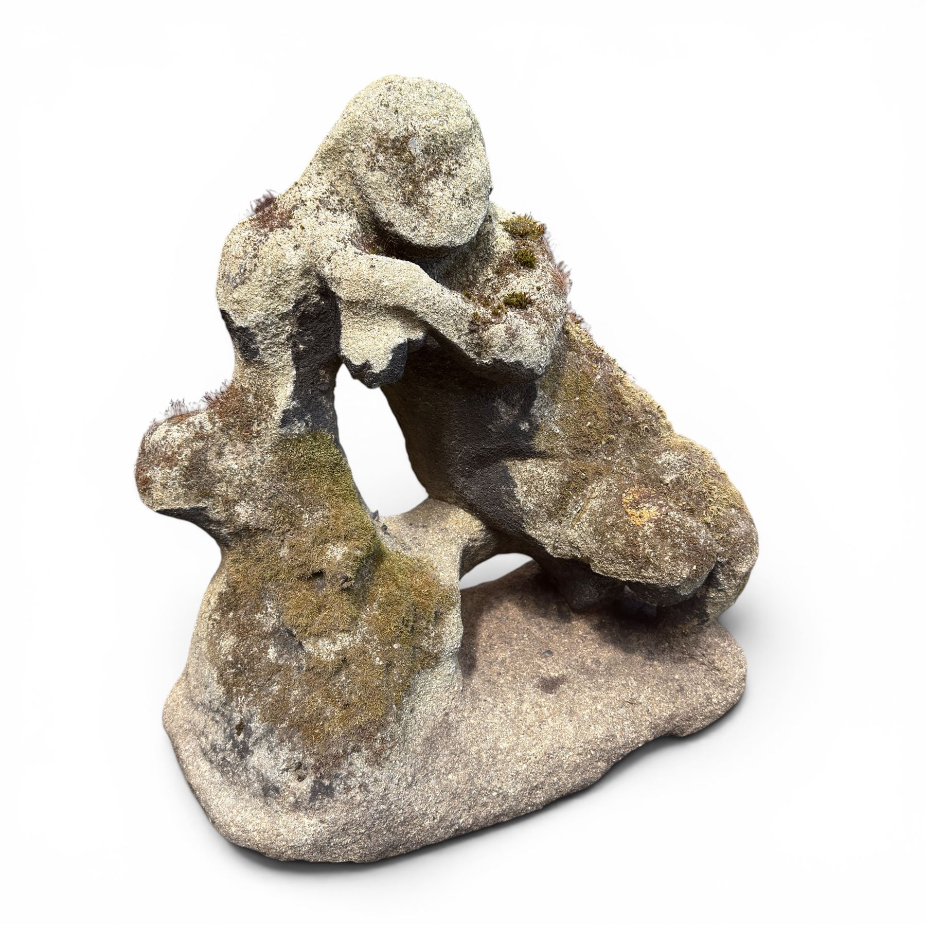 Carved composite stone garden figure, depicting a seated child with folded arms, weathered finish with moss and lichen growth, raised on an integral naturalistic base