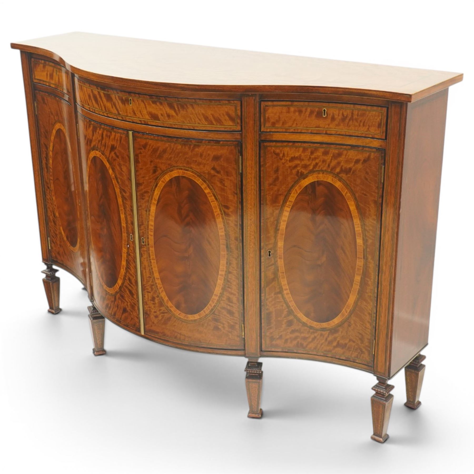 Quality handmade Sheraton Revival inlaid mahogany serpentine-front commode sideboard, shaped rectangular top with boxwood stringing, the banded frieze fitted with three drawers, over four matched-veneer cupboards with oval inlays and banding, raised on square tapering and turned feet with gadroon carving, retailed by David Hick Interiors, Jersey