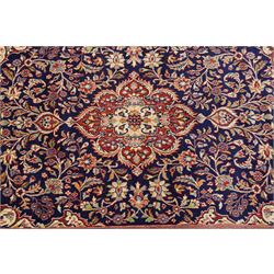 Central Persian Kashan hand-knotted rug, indigo field with central lobed medallion with pendants issuing scrolling arabesque of palmettes, flowering vines, and foliate tendrils, with conforming spandrels, with vine border and framed by guard stripes in tones indigo, ivory and madder red, with cotton fringe ends 