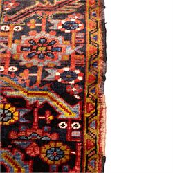 North West Persian Heriz hand-knotted carpet, madder red ground, with a large central geometric medallion with pendants, surrounded by angular foliate and floral motifs incorporating stylised rosettes, palmettes and serrated vine work, enclosed by ivory spandrels and a dark navy border with repeating geometric flowerheads and angular meandering vines, flanked by narrow patterned guard stripes, with cotton fringes
