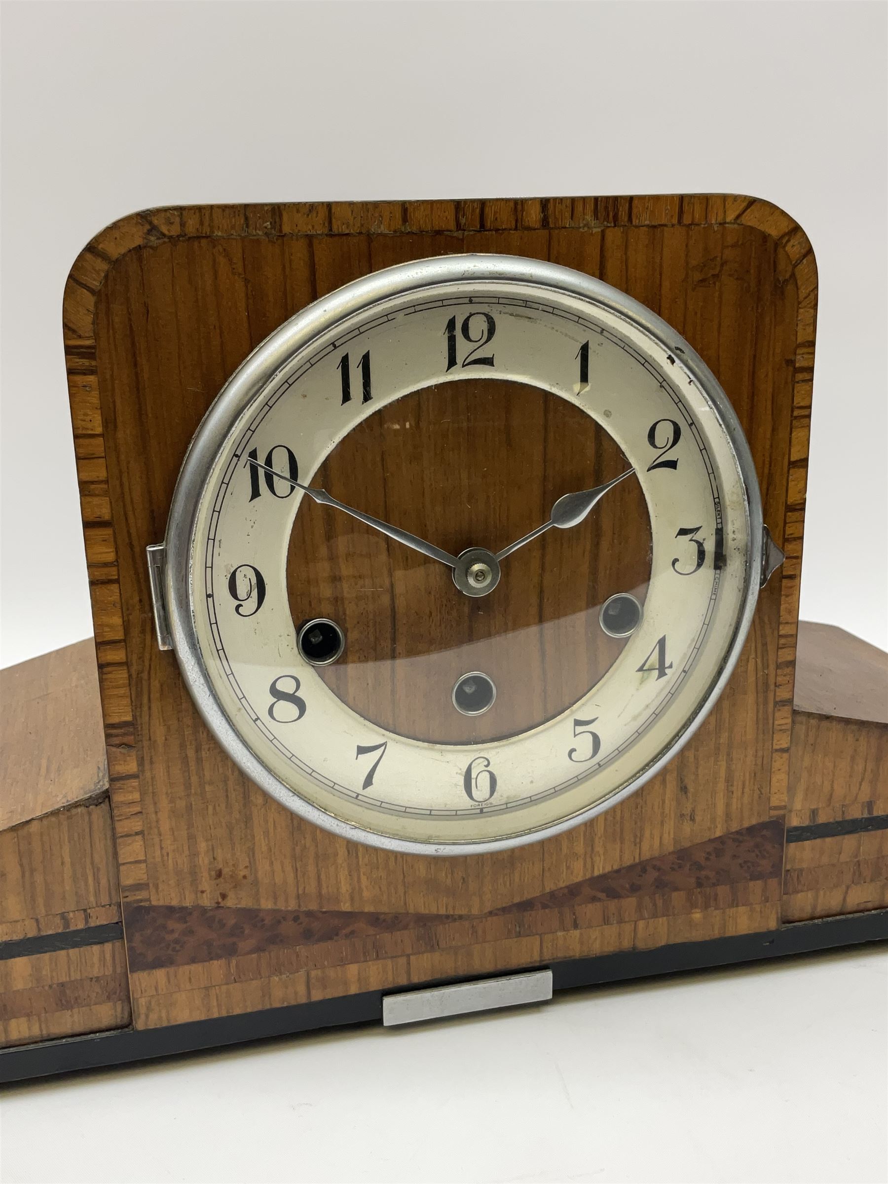 A 1930’s eight-day three train art deco Westminster chiming mantle clock with a Haller & Benzing (German) movement chiming the quarters on five underslung gong rods, silver effect chapter ring with upright Arabic numerals and minute track, strike/silent lever, chrome spun bezel with a convex glass, pierced chrome hands, walnut veneered case inlaid with cross banding and inlaid with amboyna and ebony.
With pendulum.
Haller & Benzing were a short-lived firm of German clockmakers with a factory in Villingen-Schwenningen from 1918 to 1929, mainly known for producing industrial clocks, their domestic clocks are not as well known. 
