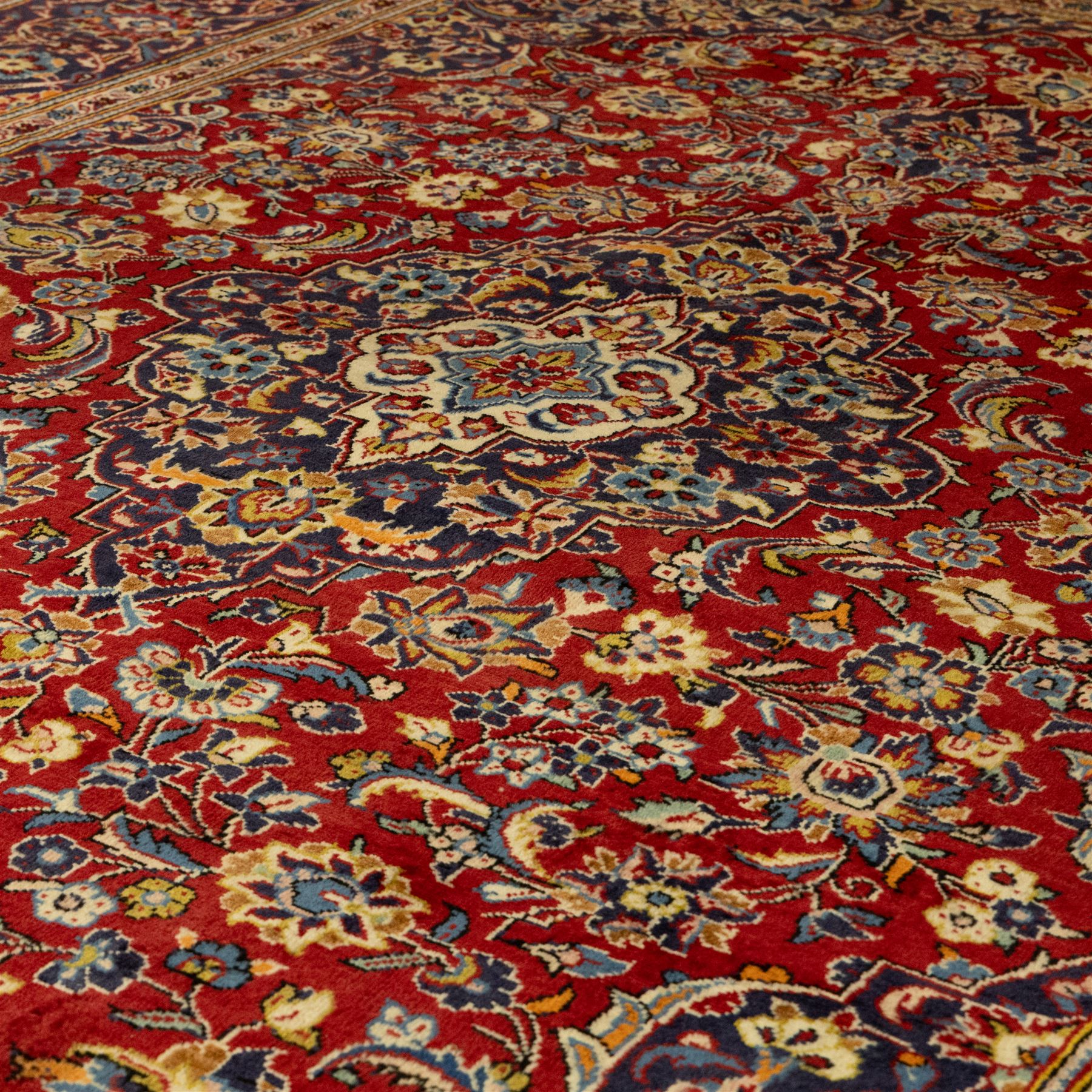Central Persian Kashan hand-knotted carpet, crimson red central field with central floral medallion with floral vine-work and palmettes, within dense floral spandrels and a wide navy blue border decorated with continuous foliate motifs and multiple guard borders, with cotton fringe ends  