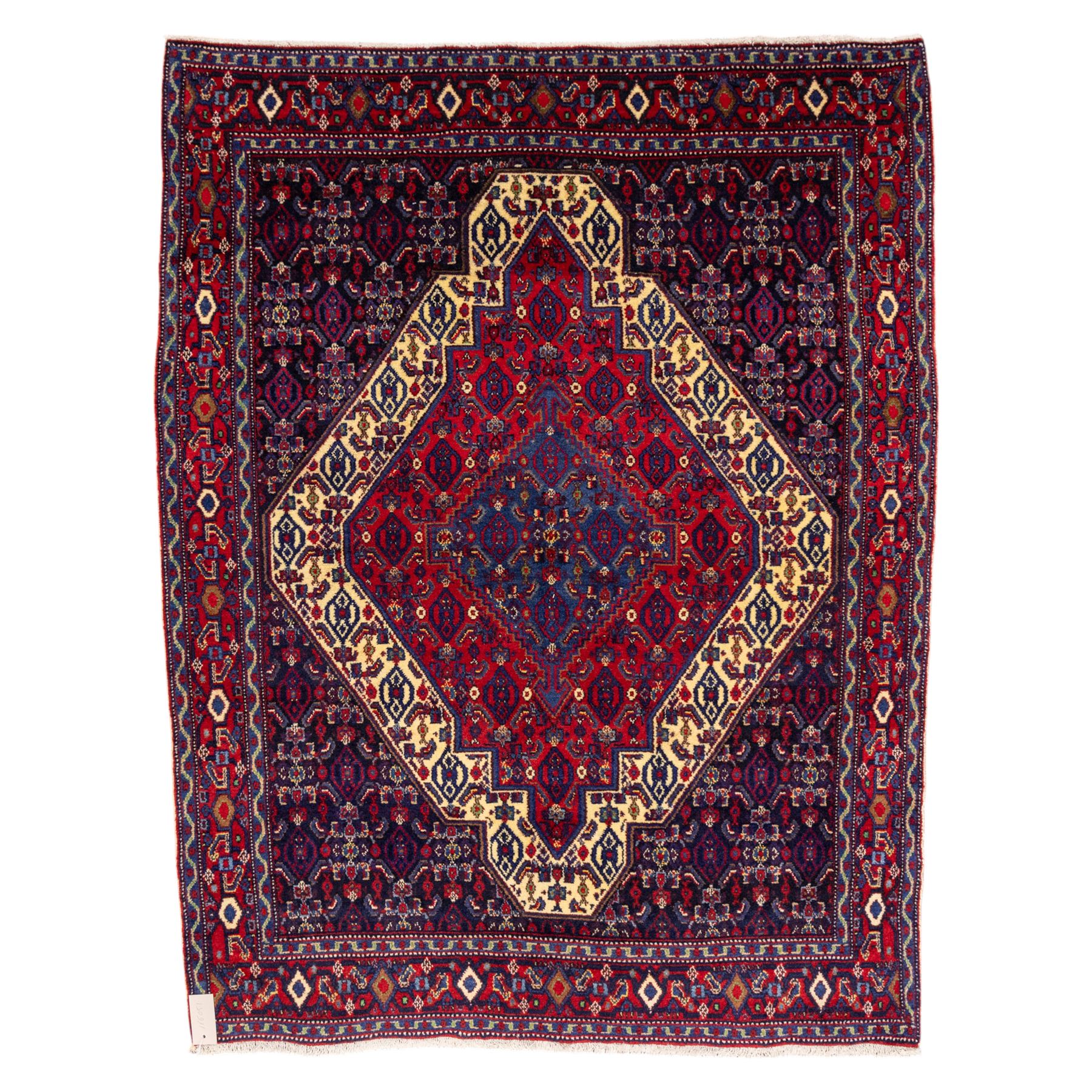 North West Persian Senneh hand-knotted rug, dark indigo field with a central bold stepped lozenge medallion of madder red and deep blue, enclosed within an ivory serrated contour forming a cruciform outline, medallion with repeating boteh and floral devices, field decorated with an all-over Herati pattern of rosettes enclosed by angular leaves principal border of madder red ground decorated with alternating floral heads, flanked by narrow guard stripes, with cotton fringe ends 