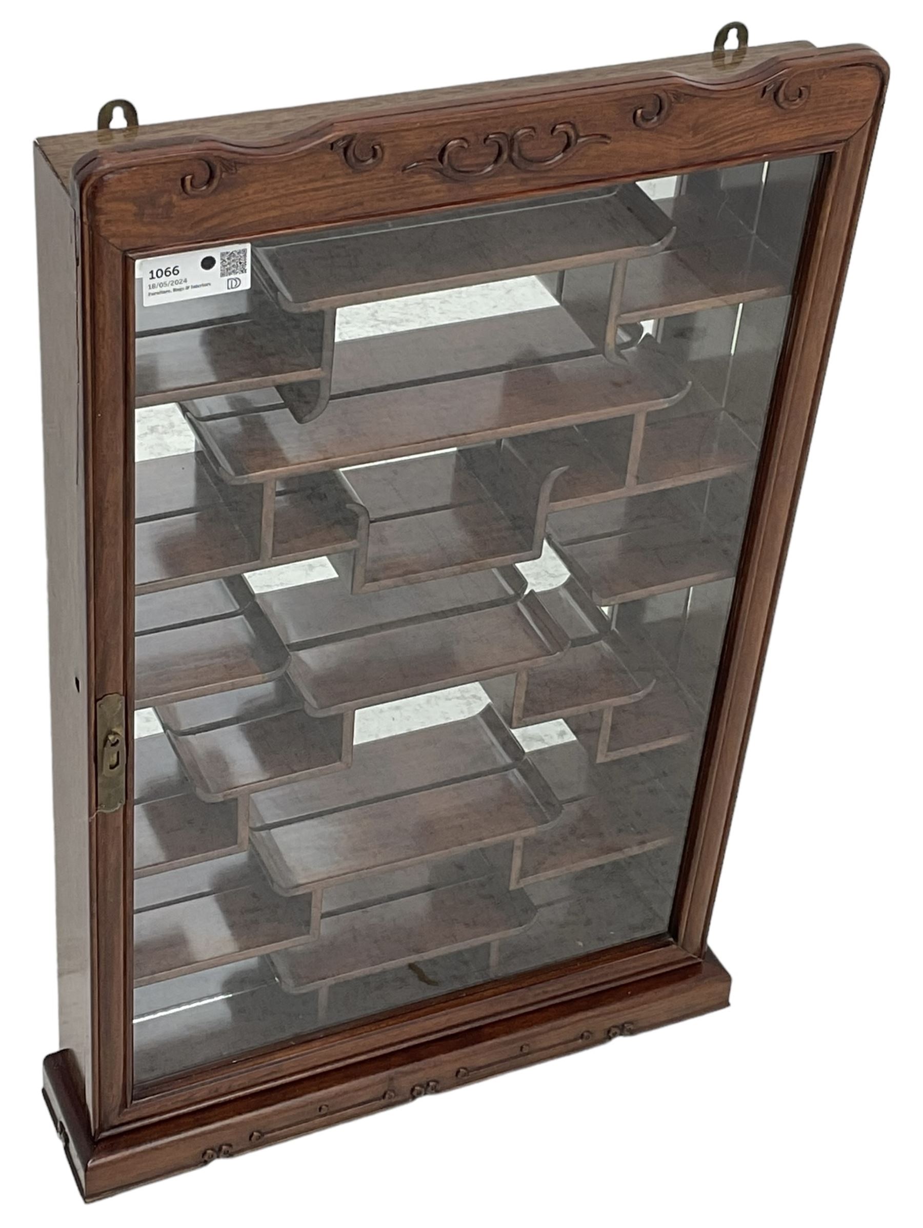 Chinese hardwood wall hanging curio cabinet, the mirror interior fitted with stepped shelves, enclosed by single glazed doors, decorated with carved scrolling foliage 