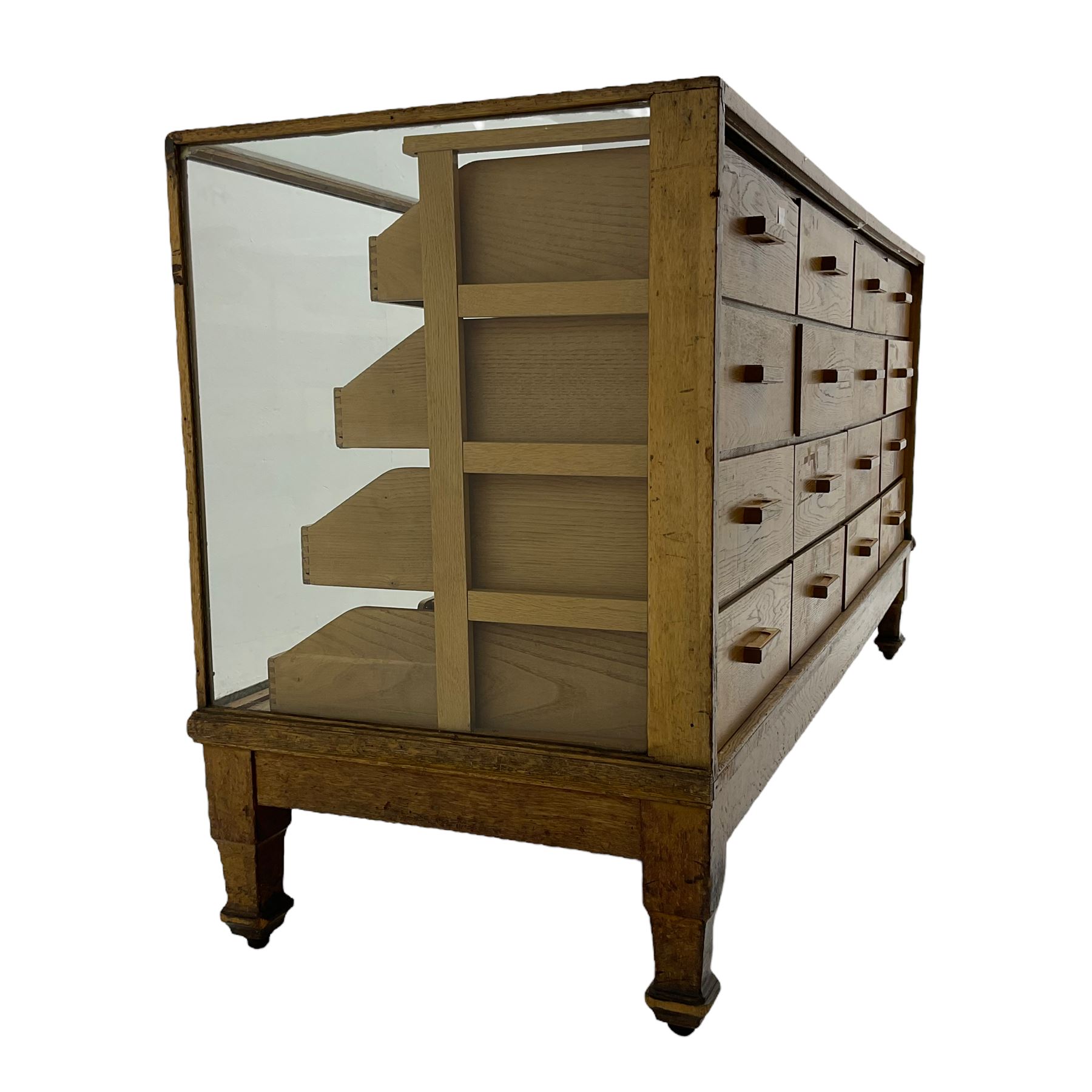 Early 20th century oak framed haberdashery shop counter, glass panel exterior enclosing sixteen graduating drawers with oak fronts and handles, raised on shaped tapering supports 