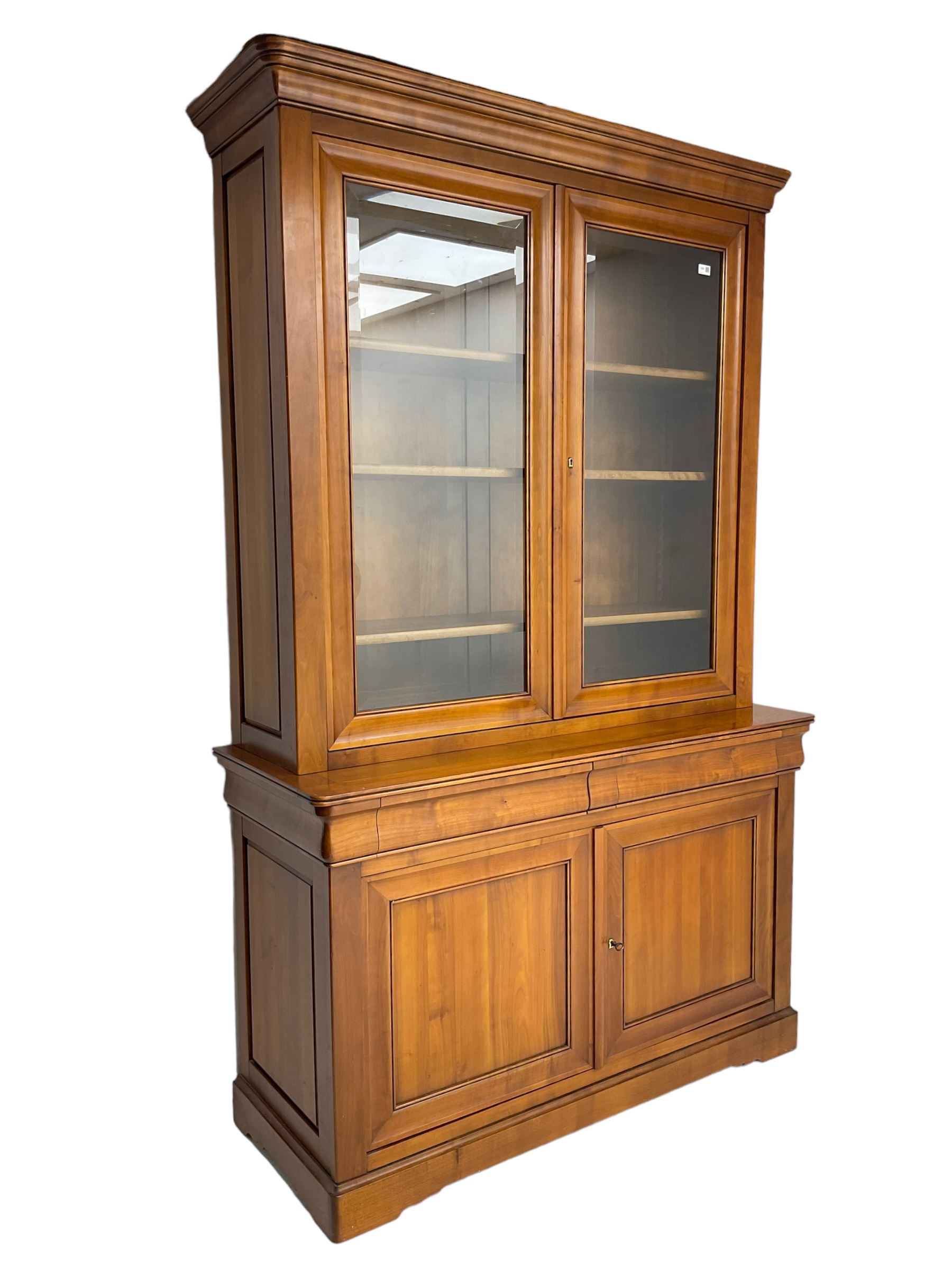 French cherry wood bookcase on cupboard, projecting cornice over two cupboard doors with bevelled glass panels enclosing three adjustable shelves, base fitted with two cushion drawers over double cupboard