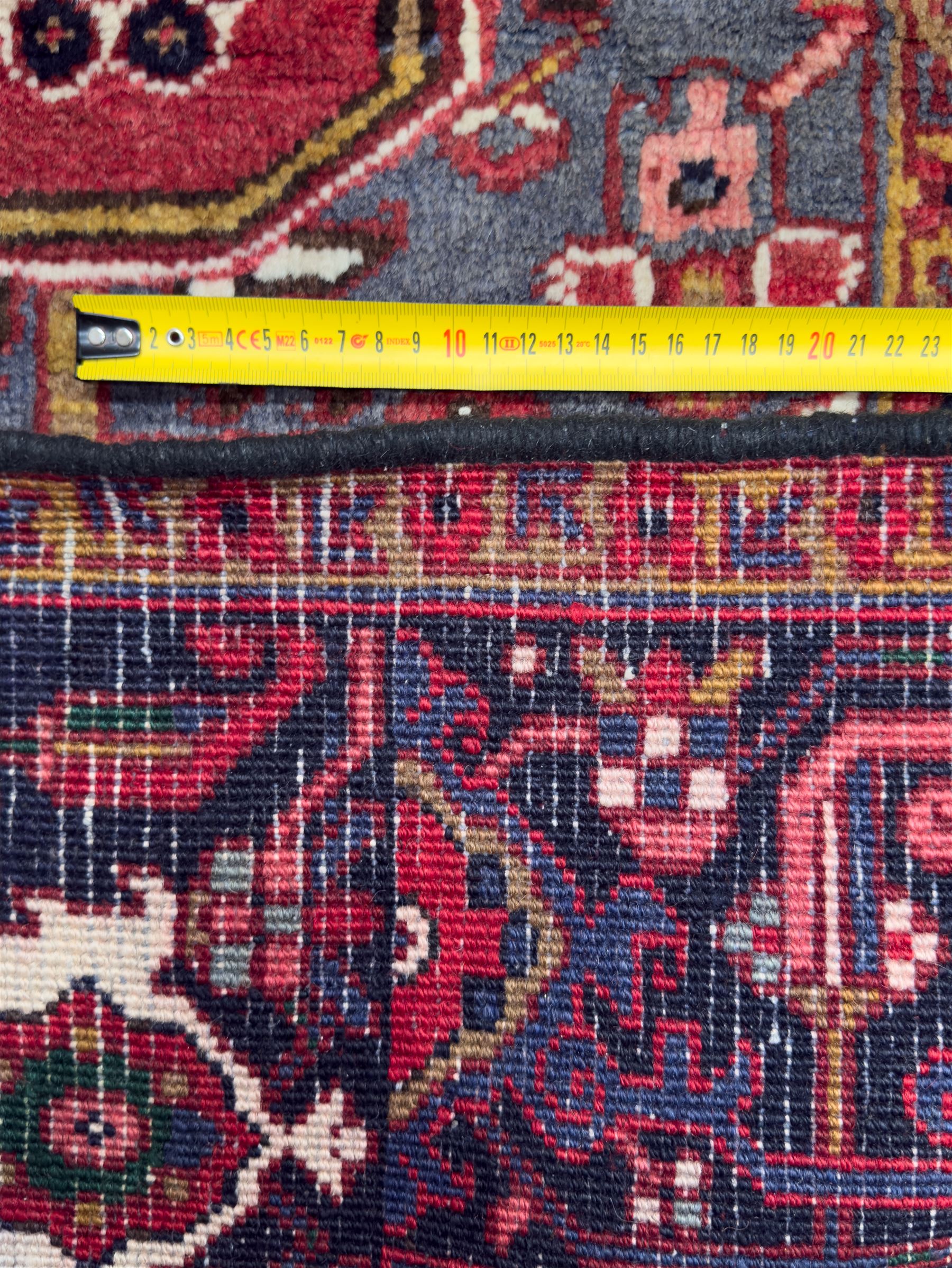 Northwestern Persian Heriz hand-knotted carpet, madder red field with a bold stepped medallion in indigo, ivory, and ivory, surrounded by angular floral motifs and stylized leaves, and the spandrels set within a dark border of rosettes and angular vine motifs, 335cmx252cm