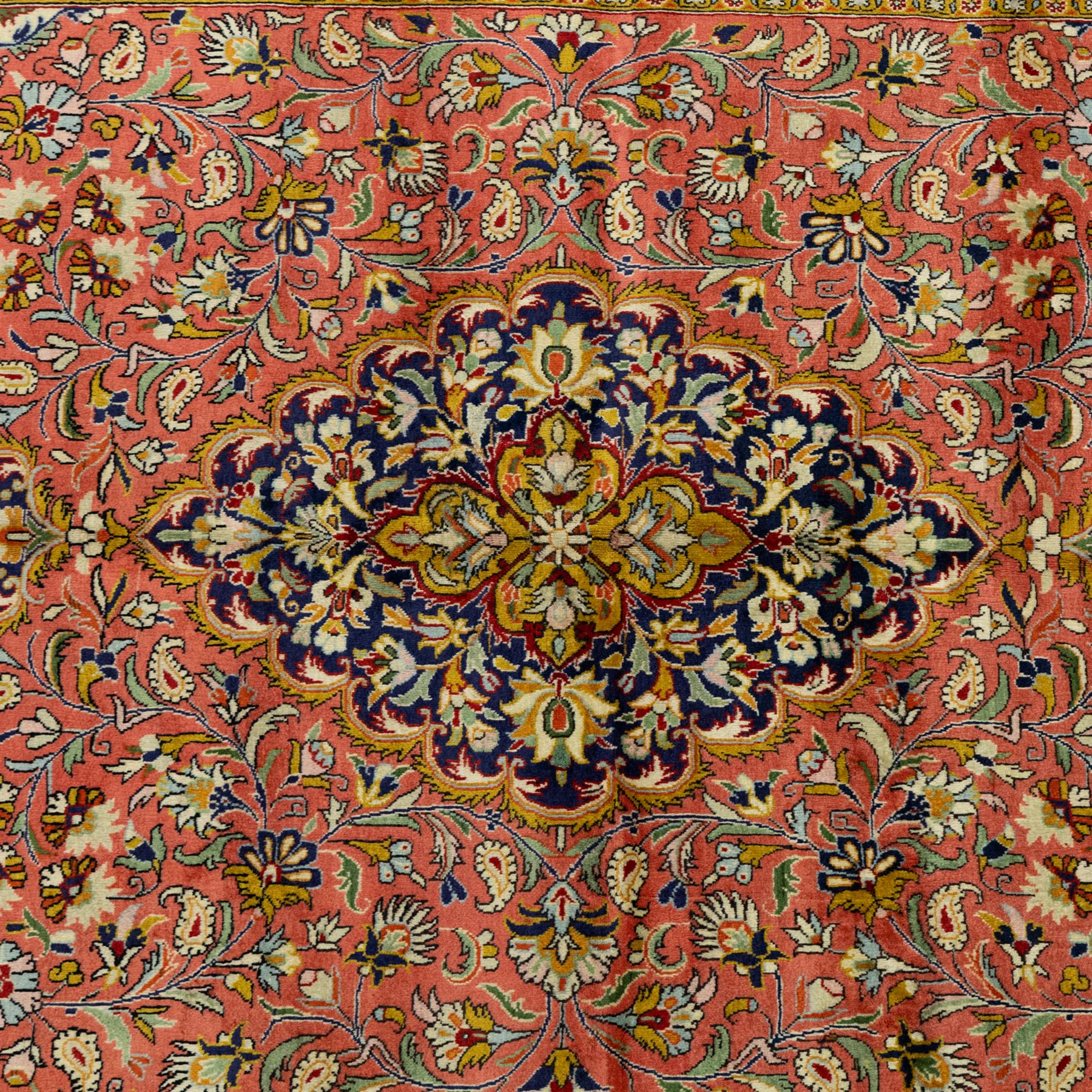 Central Persian Qom hand-knotted carpet, salmon pink ground, with a central dark navy shaped floral medallion with pendants, surrounded by dense scrolling foliate and floral motifs incorporating rosettes, palmettes and delicate vine work, enclosed by ivory spandrels and a dark navy border with repeating palmettes and meandering vines, flanked by narrow floral guard stripes, with cotton fringes