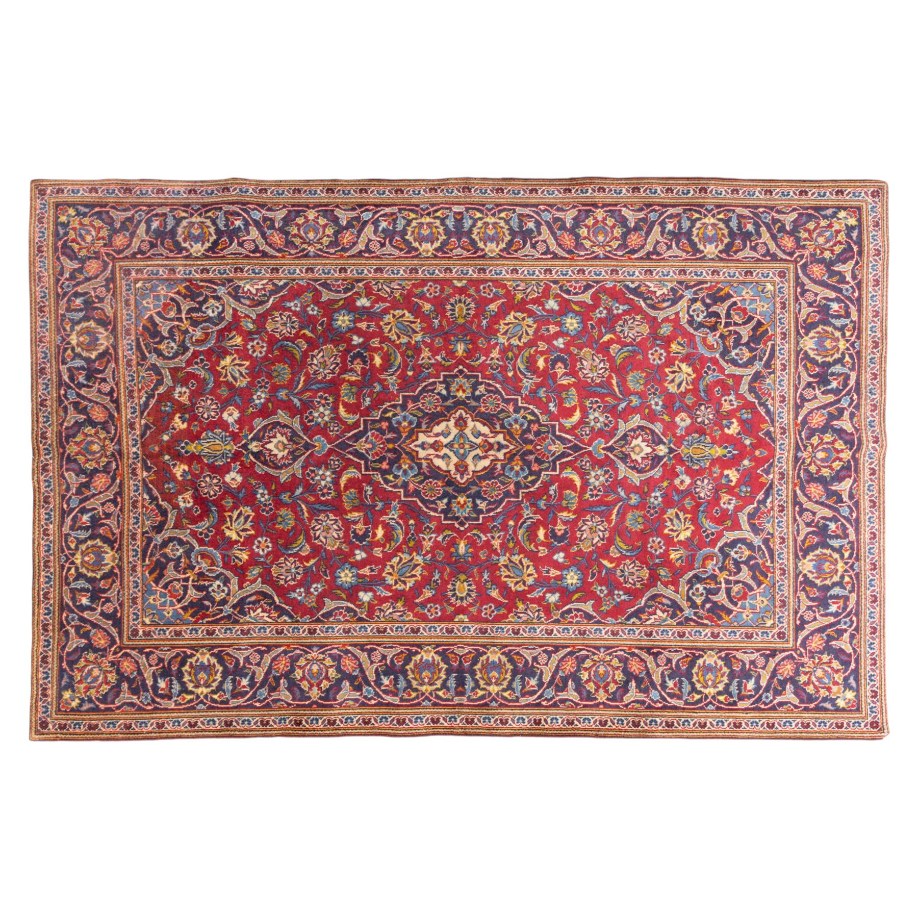 Central Persian Kashan hand-knotted rug, crimson field, profusely decorated with scrolling vines and palmettes, floral sprays around an ivory and indigo lobbed medallion enclosed by spandrels within an indigo border of arabesques between multiple guard stripes  