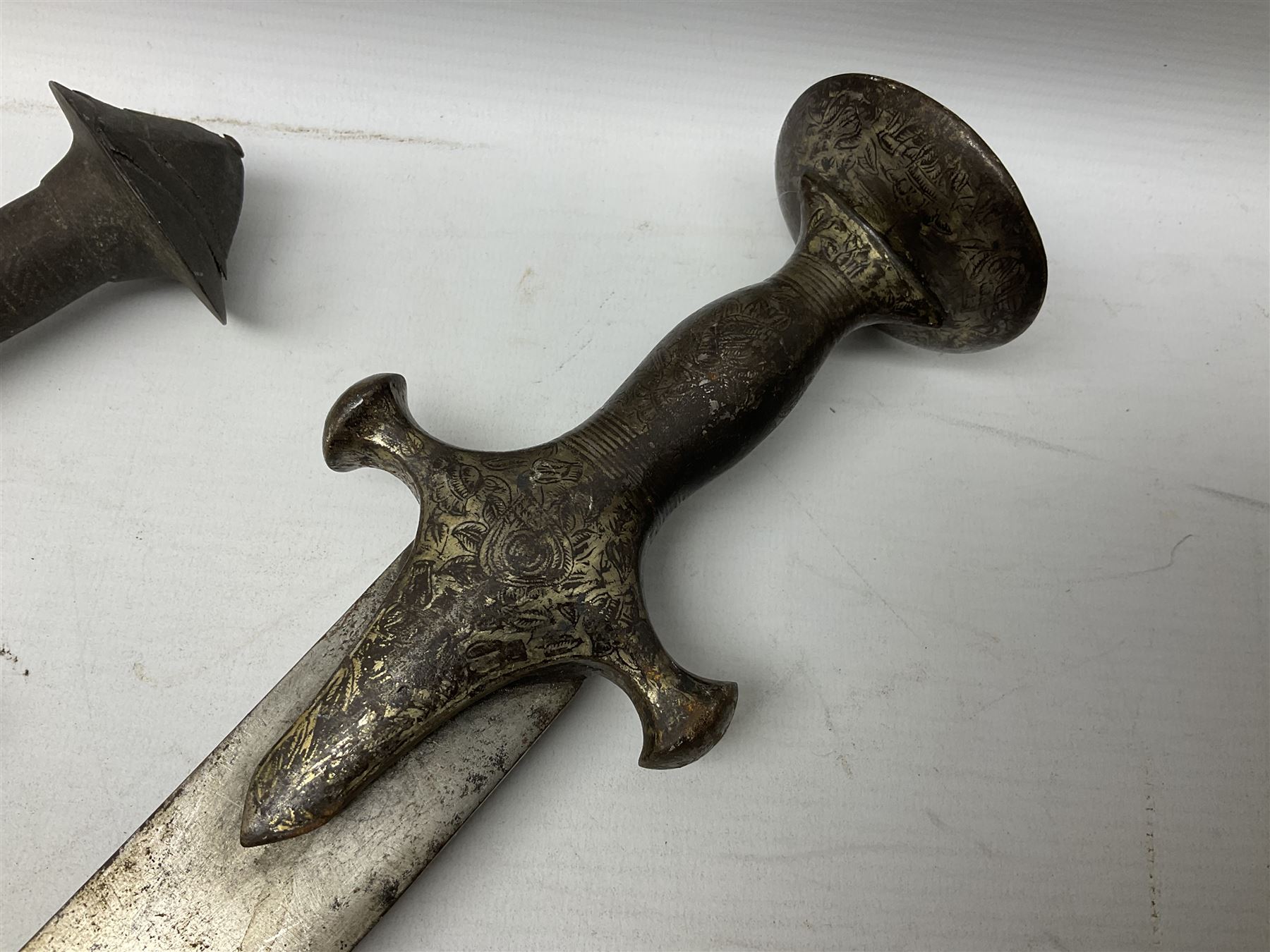 Tuareg Takoba sword with 78.5cm triple fullered blade incised with two crescent moons to either side, metal hilt with stylised floral designs to the cross-guard, decorative grip and disc form pommel cap; in tooled leather scabbard decorated with geometric designs L92cm overall; and an Indian Tulwar sword with 75cm plain curving blade and 'silver' damascened hilt (no scabbard) (2)