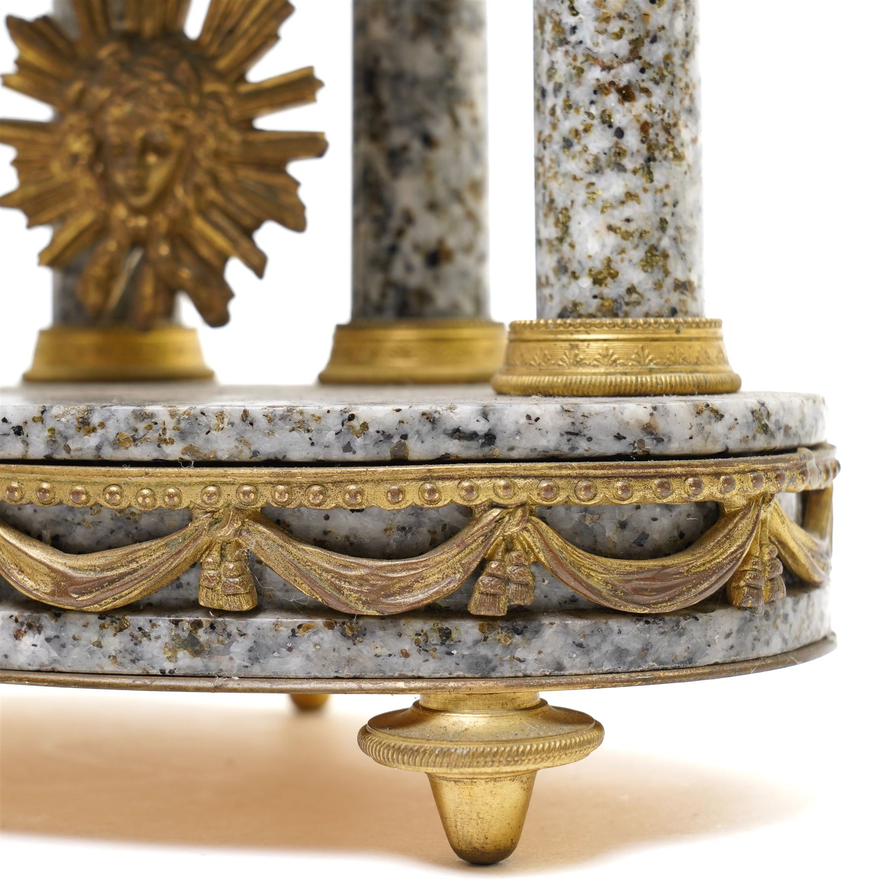 French- early 20th century portico clock garniture, 8-day drum movement decorated with laurel leaves to the glazed bezel and surmounted by a decorative urn with scroll handles, raised by four matching columns on a granite effect oval base with toupee feet, convex enamel dial with gilt pierced hands, Arabic numerals, minute markers and a garland of flowers, twin train Parisian rack striking movement, striking the hours and half hours on a bell. With a sunburst pendulum and key and a matching pair of two-light candelabra.