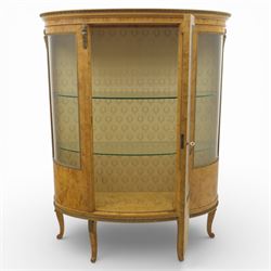 Edwardian satin walnut display cabinet, bowed oval form with moulded and dentil cornice over glazed panels, enclosing a laurel wreath damask interior with adjustable glass shelves, the frame applied with gilt metal bead mouldings and mask mounts, enclosing a shelved interior, raised on cabriole supports with moulded feet