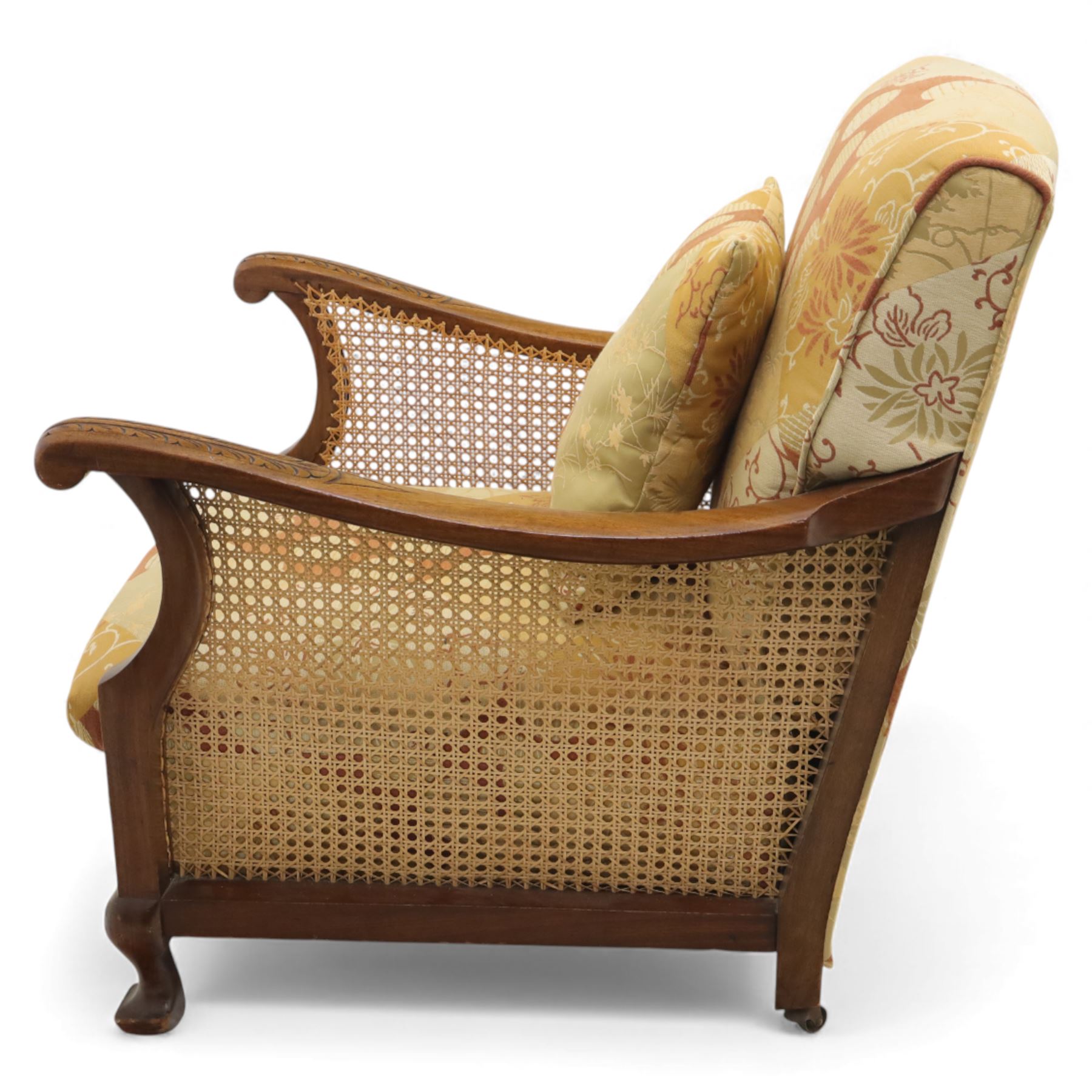 Pair of mid-20th century mahogany framed bergère armchairs, each with carved scroll arms and caned side panels, floral upholstered loose cushions, raised on cabriole front supports