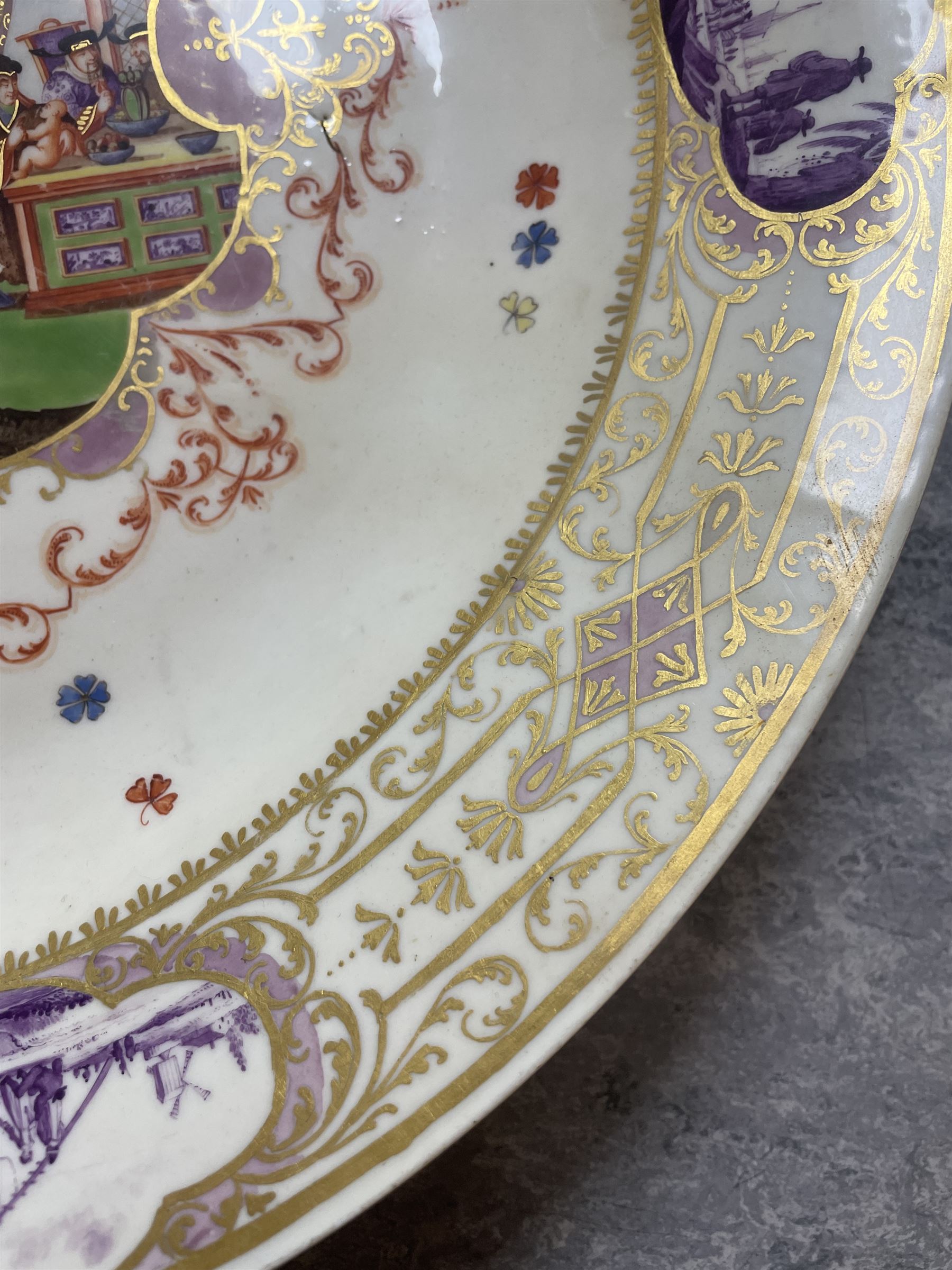 Large and impressive 18th century Meissen dish, finely painted with a chinoiserie scene to a central shaped reserve, depicting figures and attendants engaged in various pursuits, the reserve with gilt scrollwork edge, filled with Böttger lustre and detailed with iron red scrollwork, surrounded by flower heads, the rim with four quatrofoil Kauffahrtei scene panels of merchants and figures by a quayside in purple monochrome, with gilt scroll border, with under glazed blue crossed swords mark, and gilt gilders mark, D39cm
