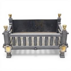 Arts and Crafts cast iron fire grate, rectangular backplate with plain upper rail over a slatted front of cylindrical tapering bars, flanked by square section uprights with fluted detail and scroll terminals, raised on stepped feet with shaped brass roundel mounts and brass finials to the corner posts