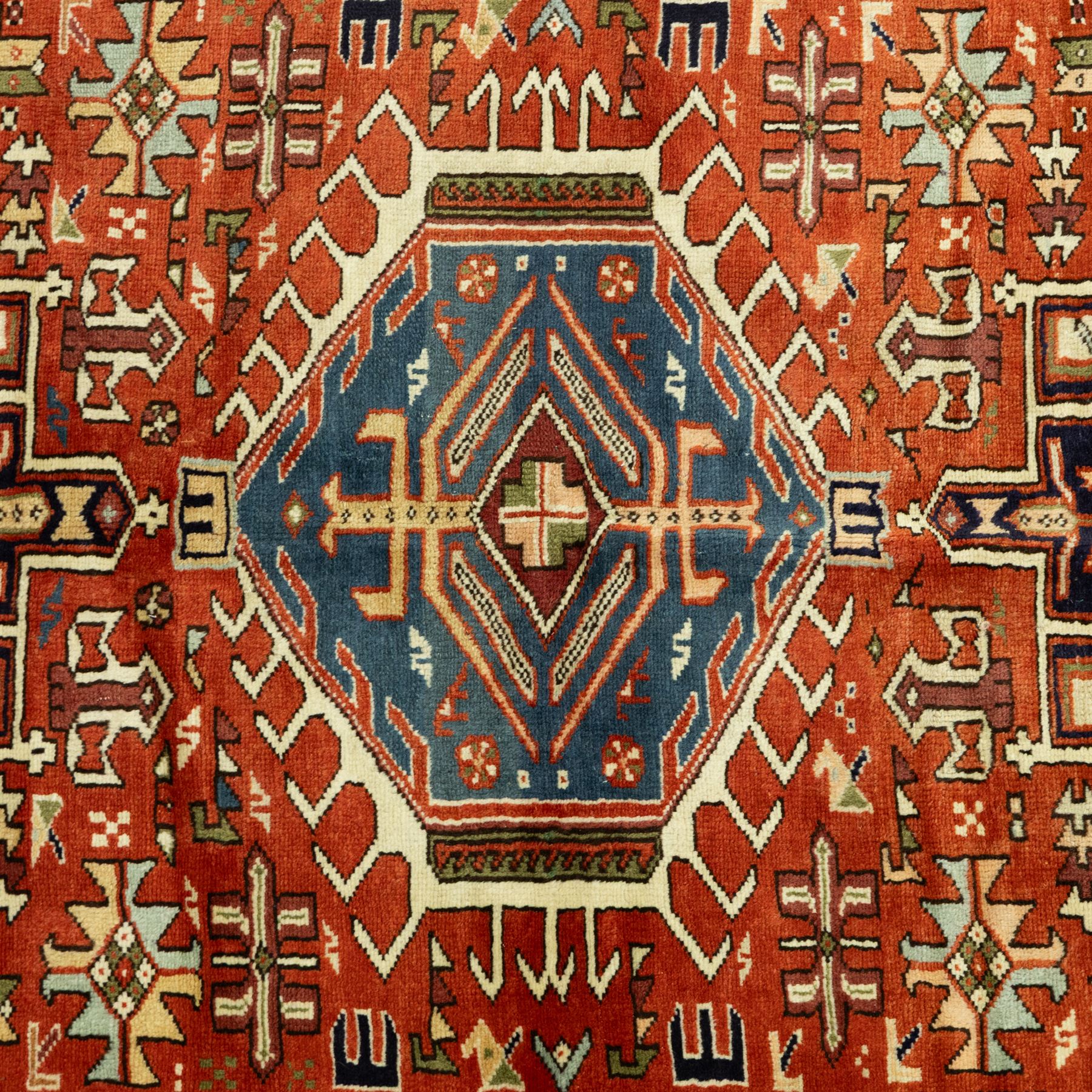 North West Persian Karajeh hand-knotted rug, terracotta ground, with a stepped geometric pole medallion design in blue, ivory and dark navy tones, surrounded by angular tribal motifs incorporating stylised flowerheads, hooked devices and geometric vine work, enclosed by a dark navy border with repeating rosettes and angular leaf motifs, flanked by narrow patterned guard stripes, with cotton fringes