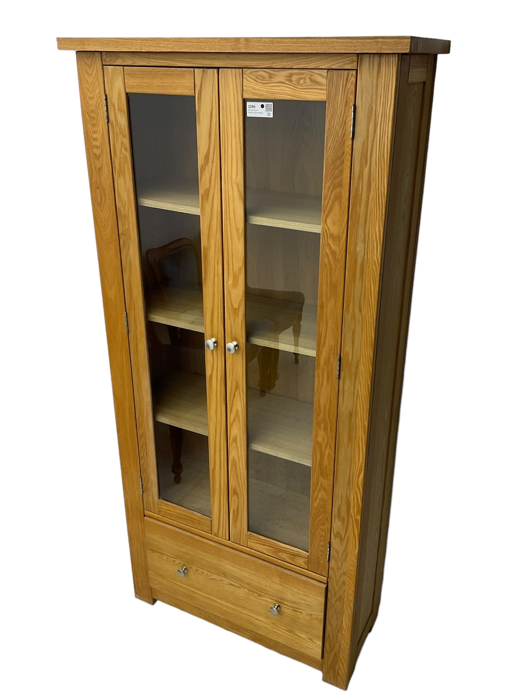 Light oak bookcase display cabinet, projecting square cornice over glazed double doors enclosing three adjustable shelves, single drawer base, on stile supports