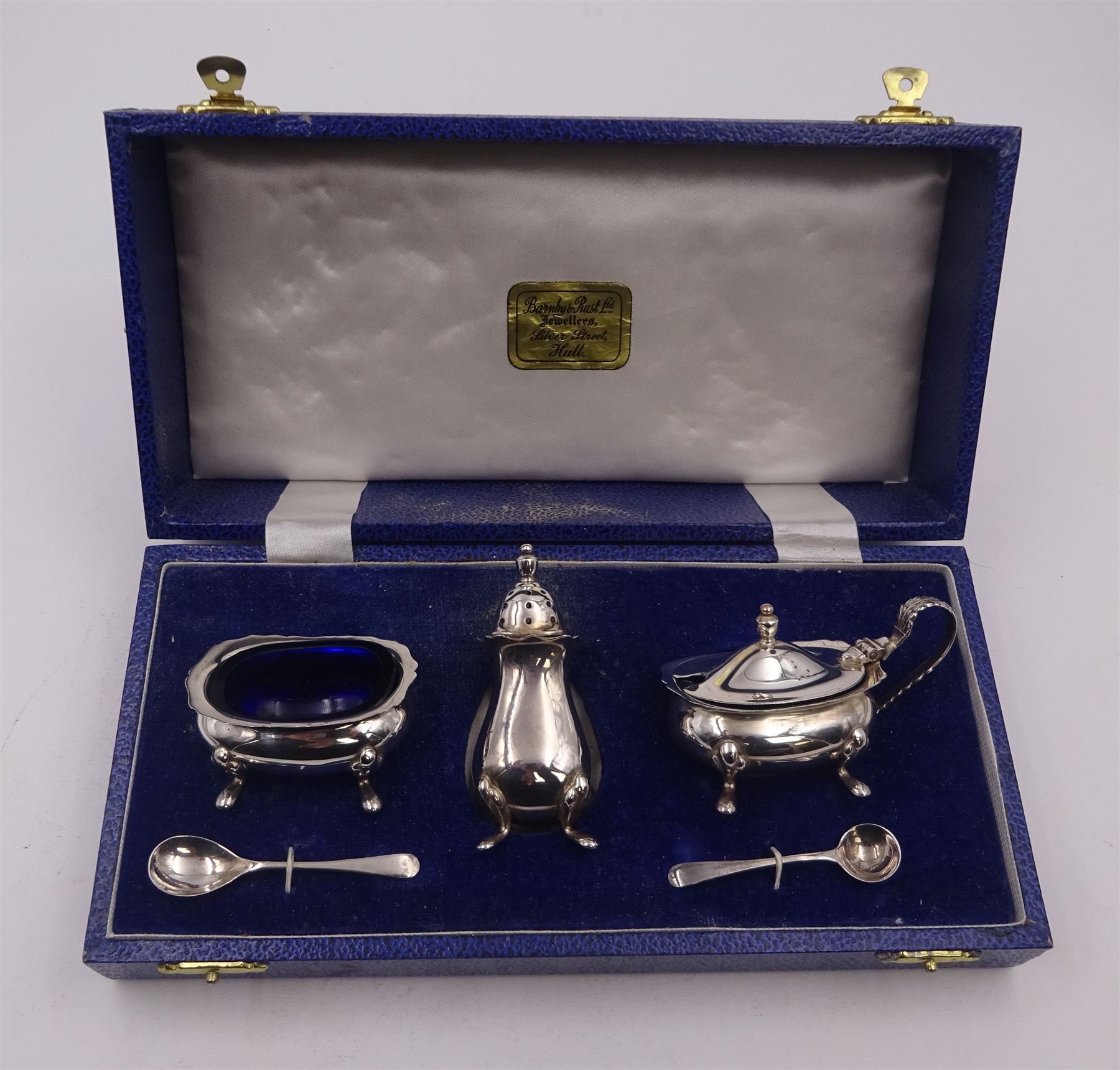 Modern three piece silver cruet set, comprising pepper shaker, open salt, mustard pot and cover, all of typical form with shaped rims and upon four pad feet, the pepper shaker and mustard pot with urn shaped finials, together with two matching condiment spoons, all hallmarked Turner & Simpson Ltd, Birmingham 1978, the mustard pot and salt with blue glass liners, all within fitted case