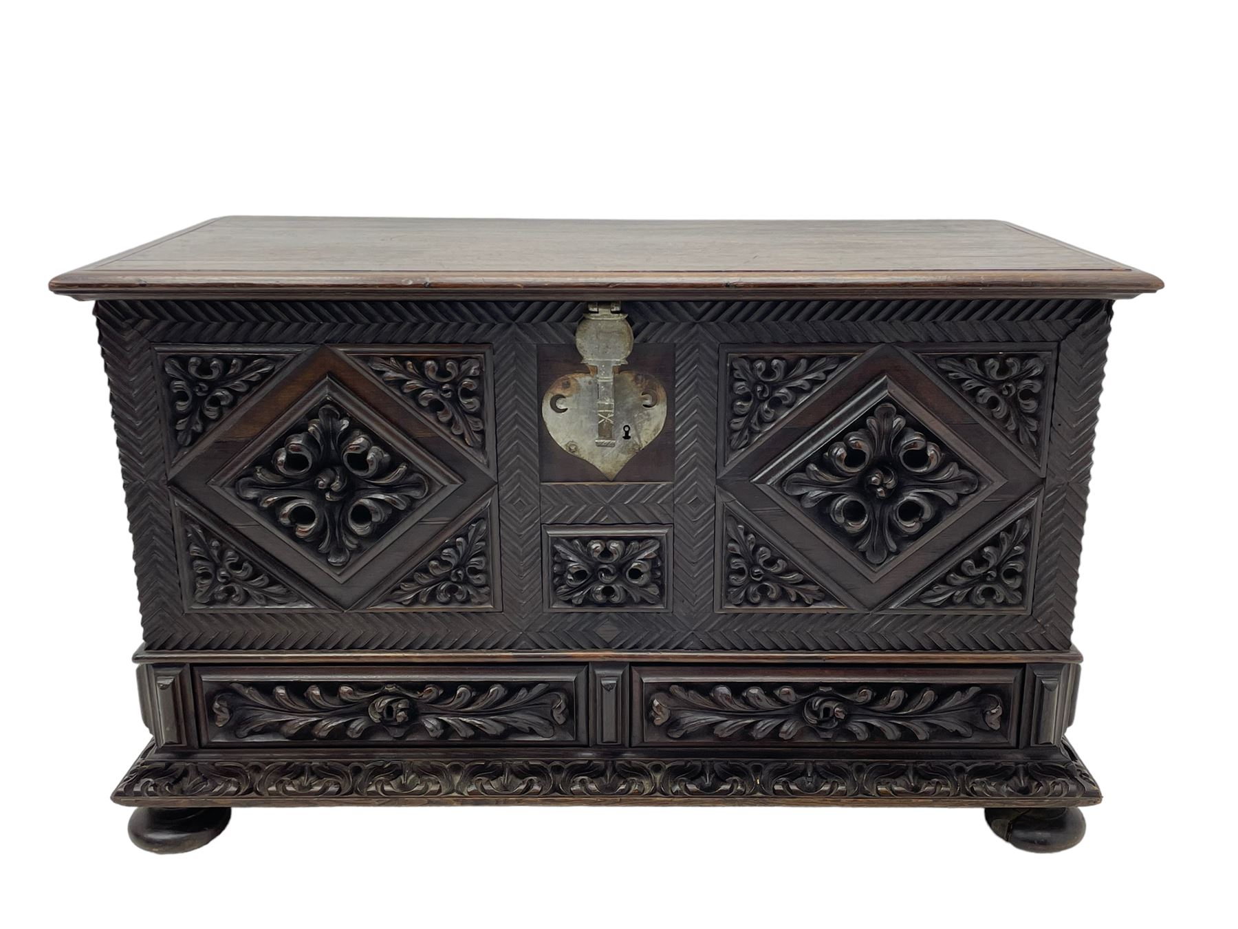 Early 20th century carved hardwood kist or mule chest of small proportions, moulded rectangular hinged lid over foliage carved geometric lozenge panels, the frame carved with chevron banding, fitted with two drawers, the drawer fronts carved with extending leaf motifs over a foliage carved lower moulding, on turned feet