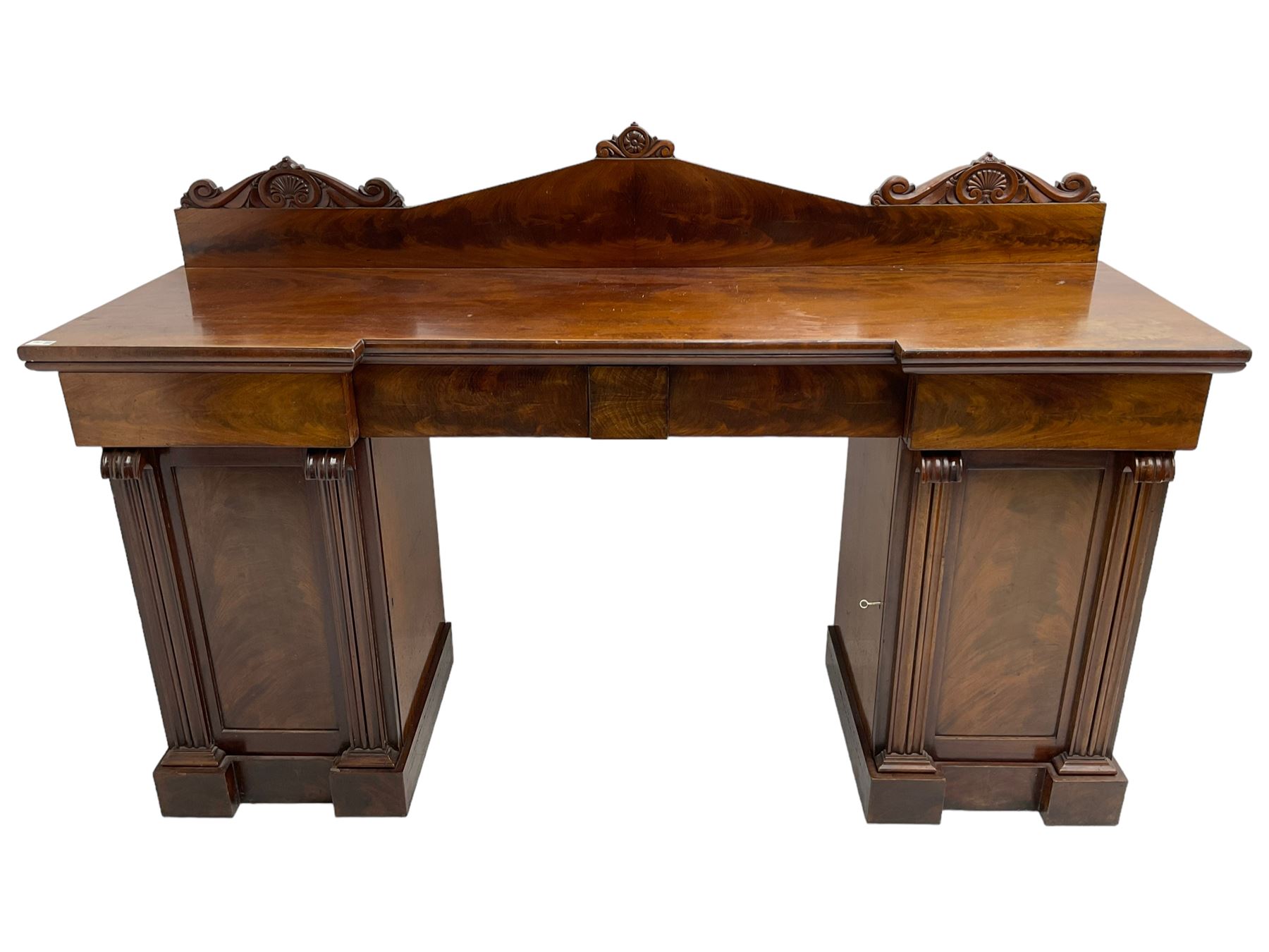Early 19th century mahogany reverse break-front twin pedestal sideboard, raised back carved with cartouche and rosette detail with extending scrolling, fitted with two frieze drawers, the flanking panelled cupboards enclosing sliding trays and cellarette drawer