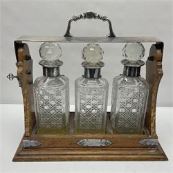 Silver plate mounted oak Tantalus, with three square glass decanters each with silver plate collars, one hallmarked silver decanter label, complete with key, H36cm