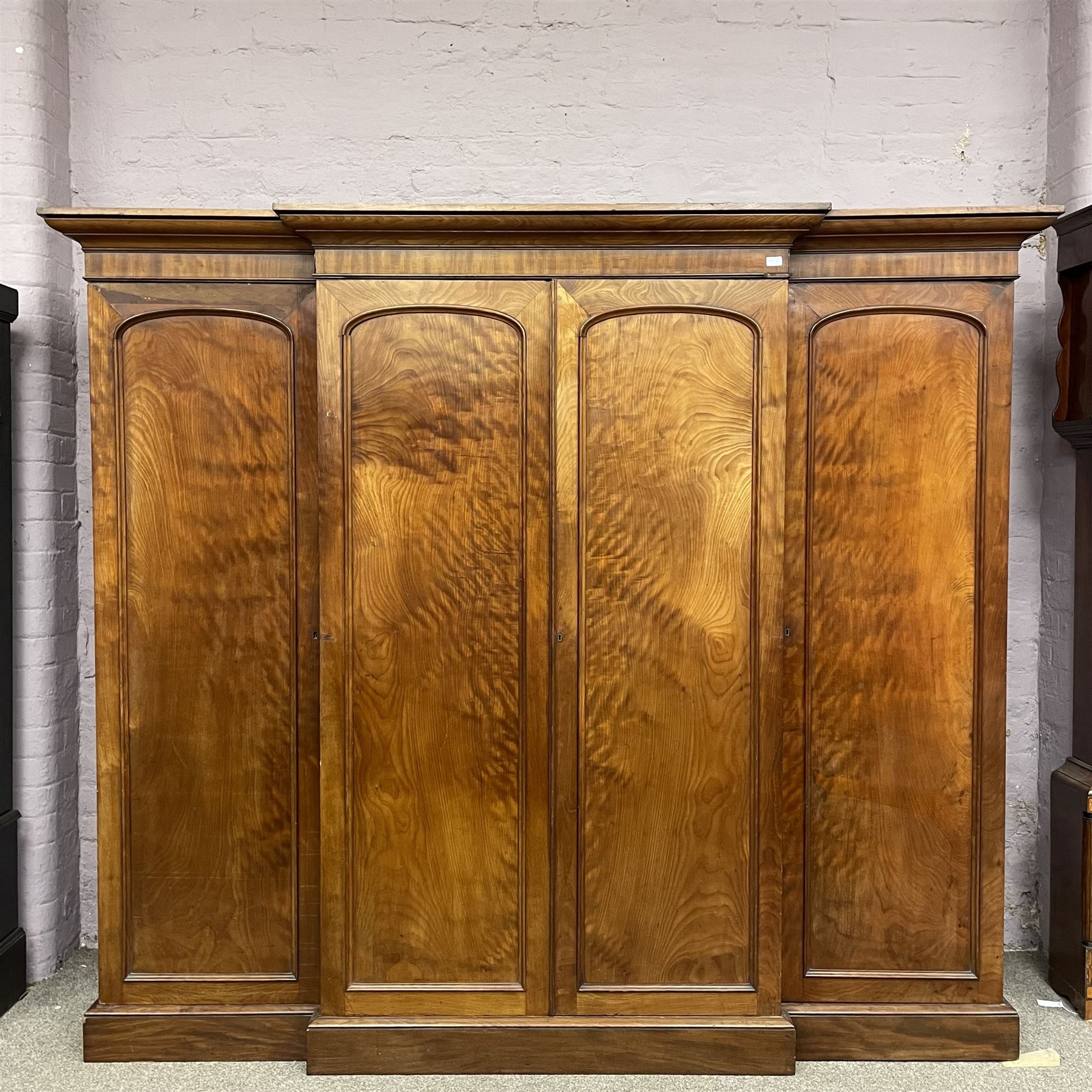 19th century figured mahogany quadruple breakfront press wardrobe, projecting cornice with banded frieze over arch panelled doors, the interior fitted with three sliding trays over two short and three long graduating drawers with recessed brass handles, flanked by two hanging cupboards with hooks