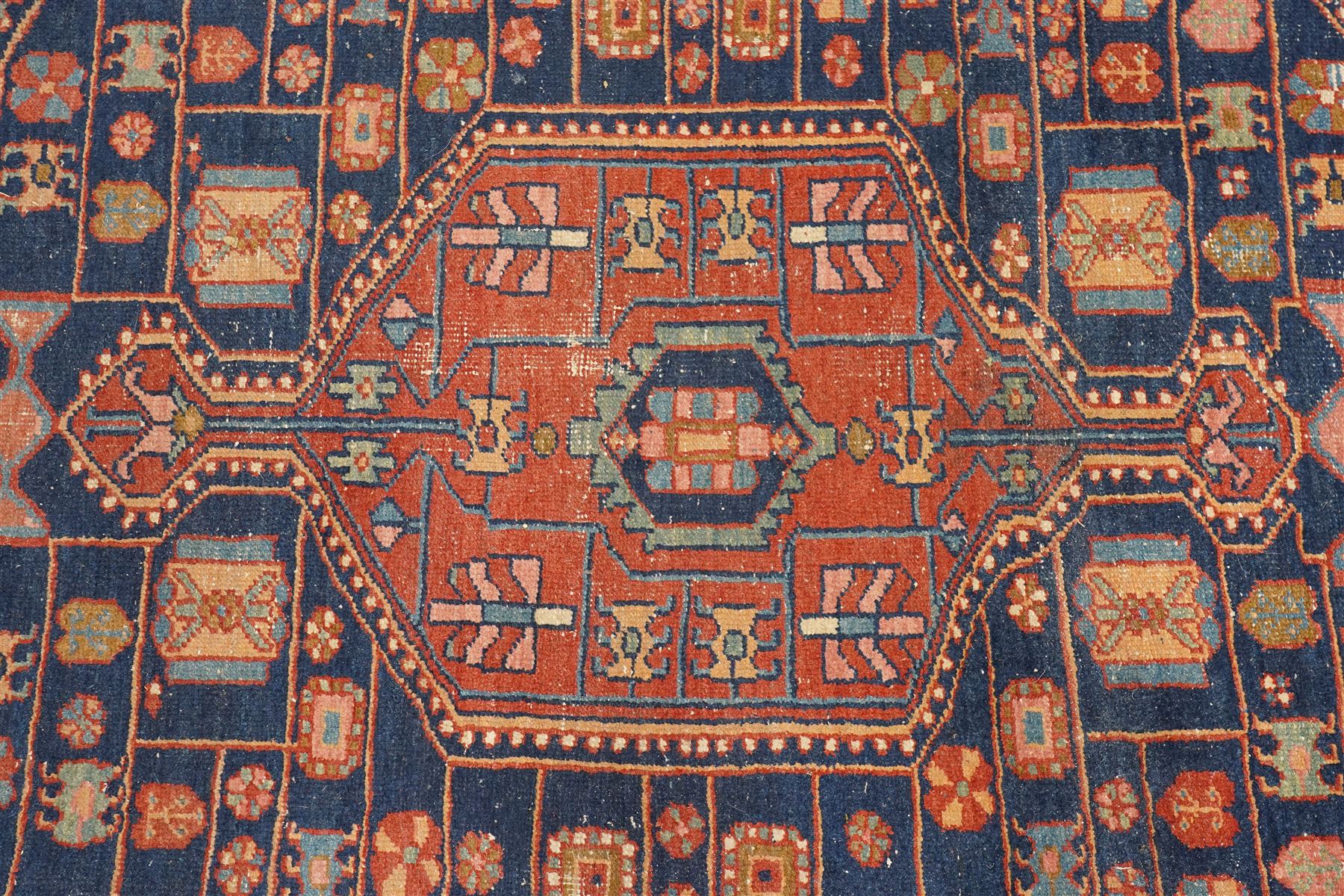 Persian Heriz hand-knotted carpet, terracotta ground centred by large dark navy geometric medallion with stepped outline, decorated throughout with stylised flowerheads, palmettes and geometric motifs in blue, green and ochre, enclosed by diagonal geometric border with narrow guard stripes