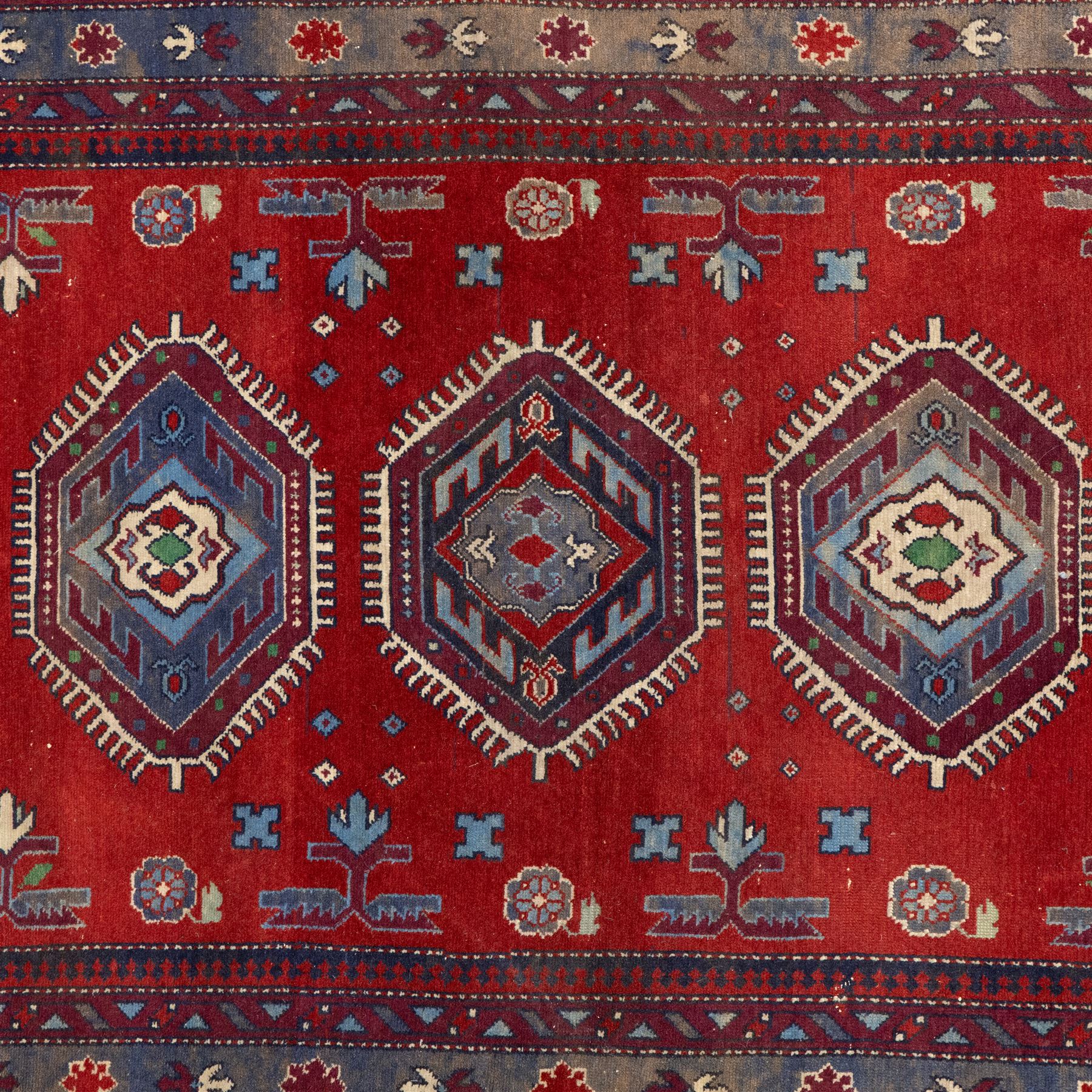 Caucasian Kazak rug, red field decorated with a column of stepped hexagonal medallions in blue, ivory, and crimson, flanked by stylized floral and geometric motifs, enclosed by multiple borders of rosettes and hooked designs 
