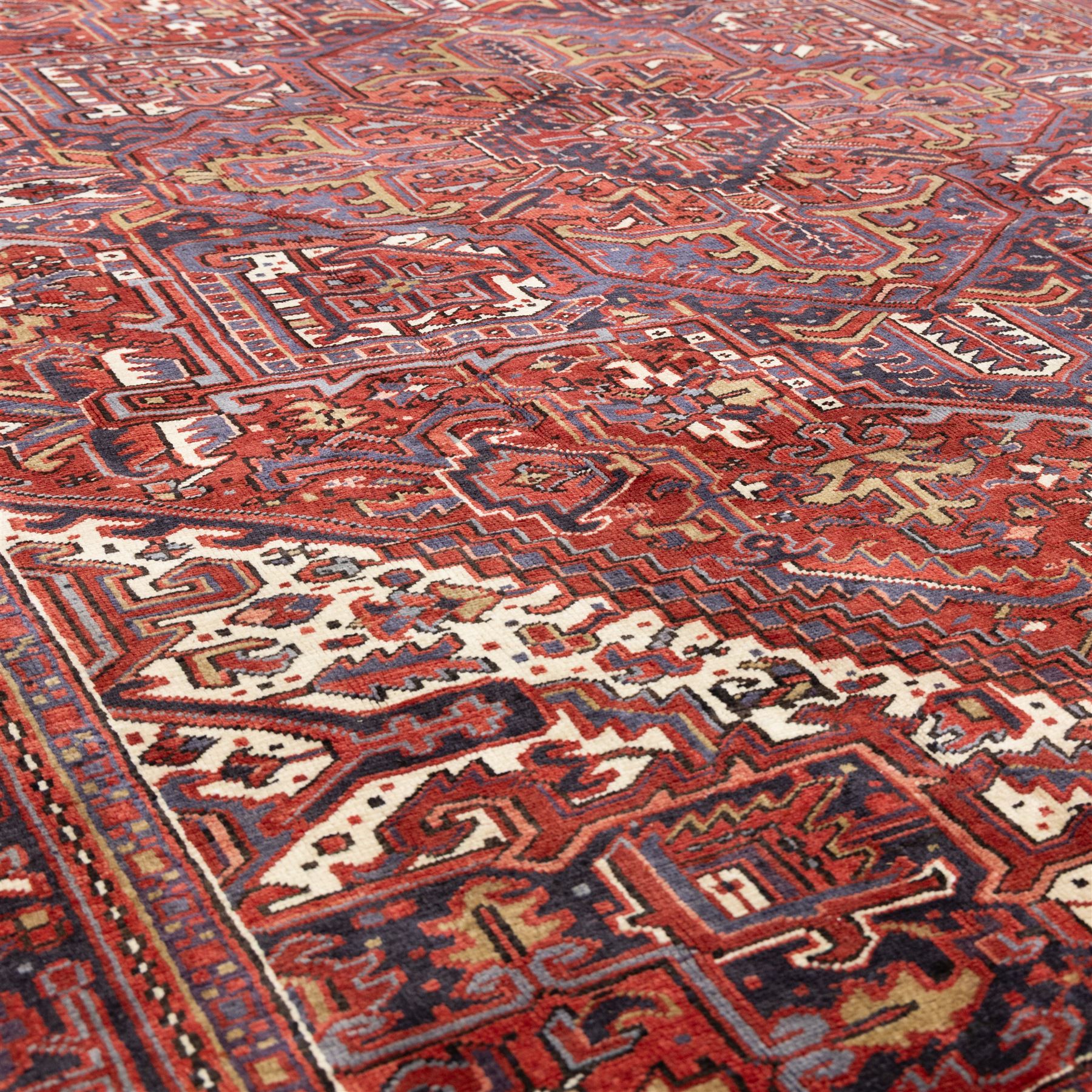 North West Persian Heriz hand-knotted carpet, madder red field with central bold angular steeped medallion with serrated outlines, flanked by stylized palmettes, hooked leaves and geometric motifs, spandrels similarly decorated within a wide geometric border and multiple guard borders in tones of madder red, indigo blue, ivory and olive green, with cotton fringe ends  