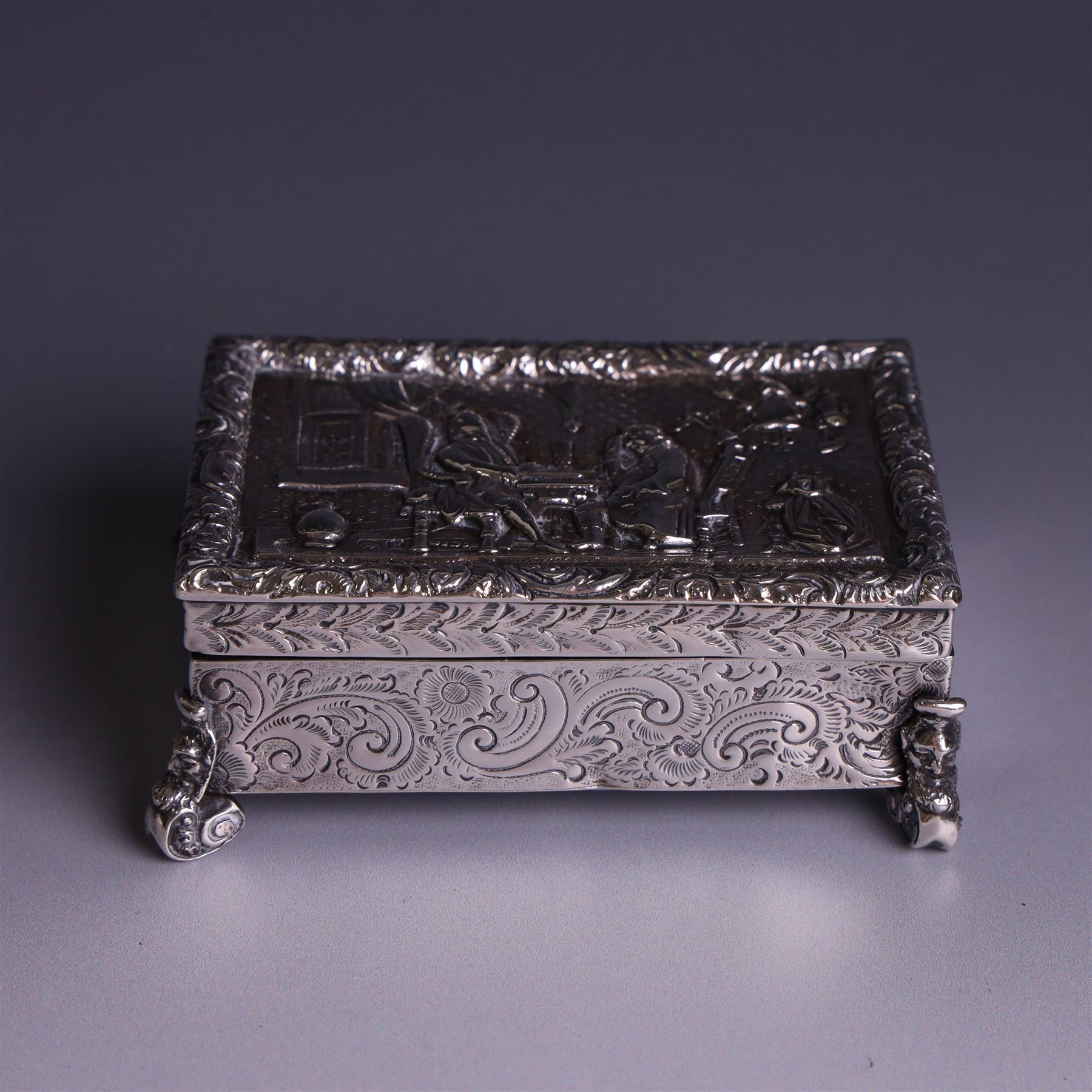 Victorian silver tobacco box, rectangular form, hinged lid embossed with a gaming scene, the body engraved with scrolls and flowers, upon four scrolling mask feet, hallmarked George Heath London 1885, H4.5cm, W10.5cm