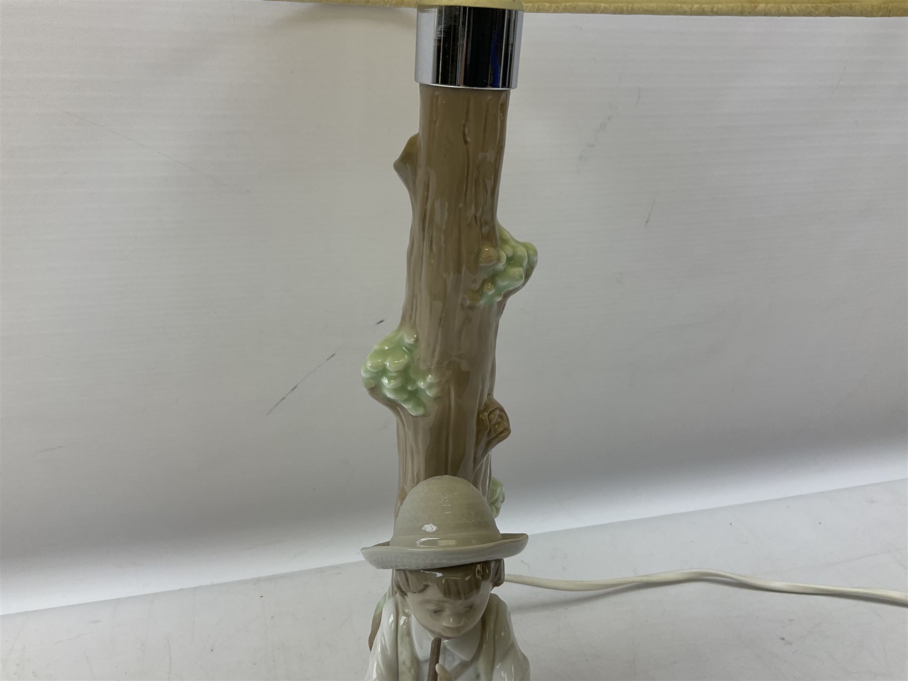 Nao table lamp modelled as a boy playing the flute upon logs with two recumbent sheep at his feet, the stem modelled as a tree trunk, with fabric shade, with printed mark beneath, H46cm