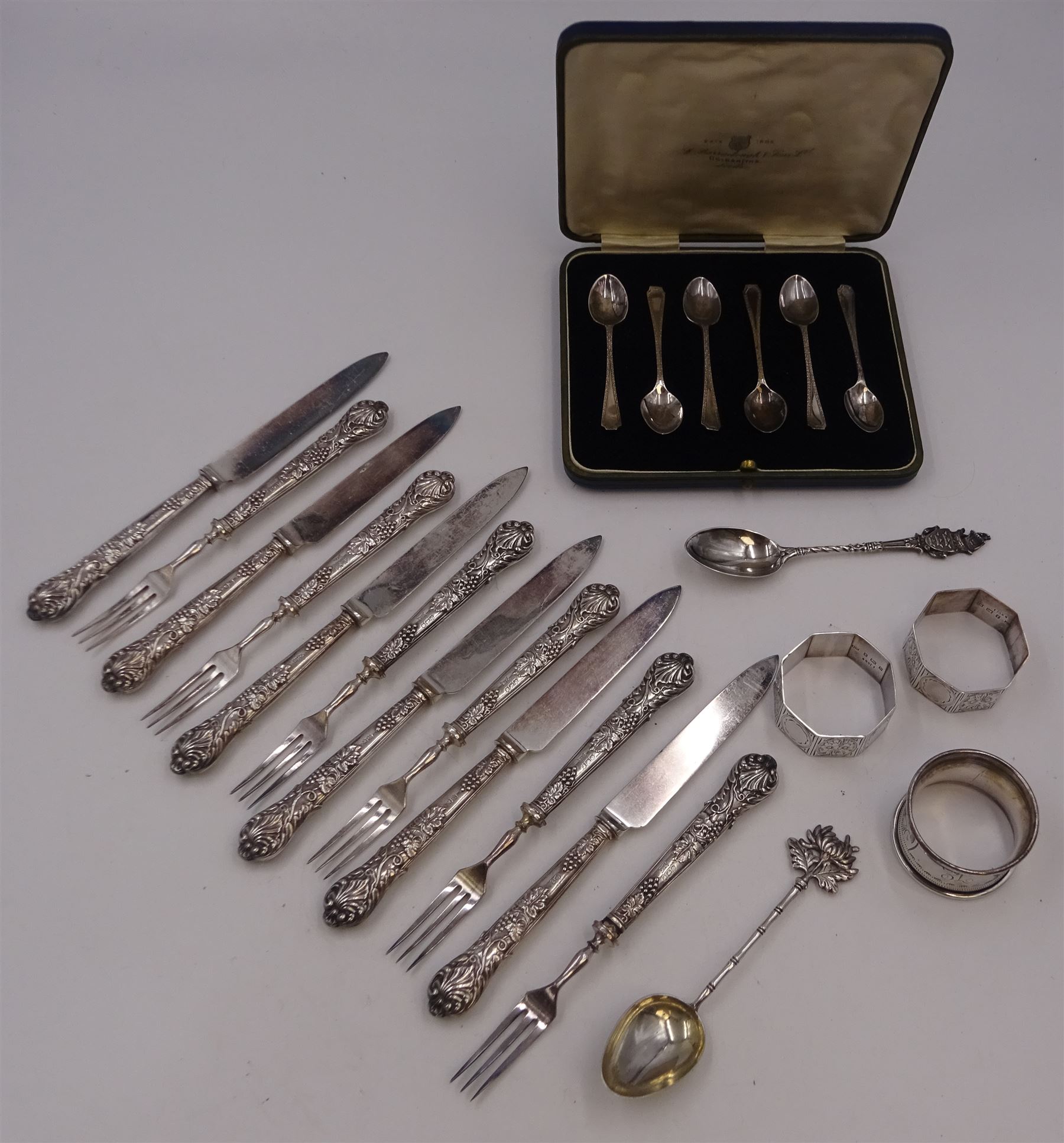 Set of 1930s silver handled fruit knives and forks, for six place settings, the handles decorated in relief with fruiting vines, hallmarked Walker & Hall, Sheffield 1934, together with a set of six 1920s silver egg spoons, with pin prick engraving to handle, hallmarked Thomas Bradbury & Sons Ltd, Sheffield 1929, in fitted case, three silver napkin rings, to include a pair, of octagonal form, with floral engraving, hallmarked John Rose, Birmingham 1951 and two silver souvenir spoons