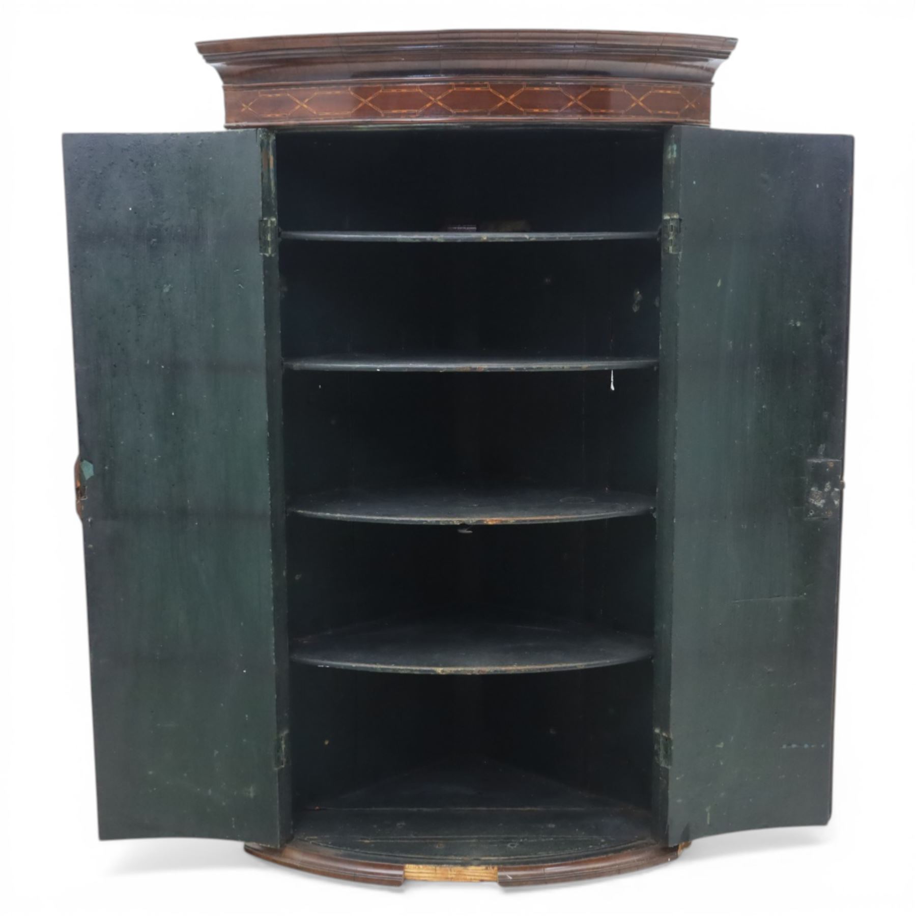 George III mahogany bow-front corner cupboard, projecting moulded cornice over satinwood inlaid frieze with geometric crossbanding, enclosed by twin shaped panel doors with line inlay and ornate brass escutcheon plates, painted interior fitted with shaped shelves, on moulded plinth base
