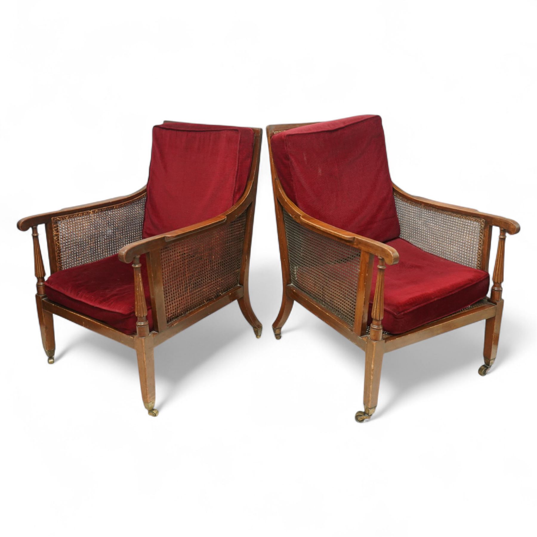 Pair of early 20th-century mahogany Bergère armchairs in the manner of Gillows, the back and seat with woven cane panels with loose red velvet cushions, the frame with swept arms with tapered and reeded uprights, raised on tapered square-section front legs terminating in brass cup castors 