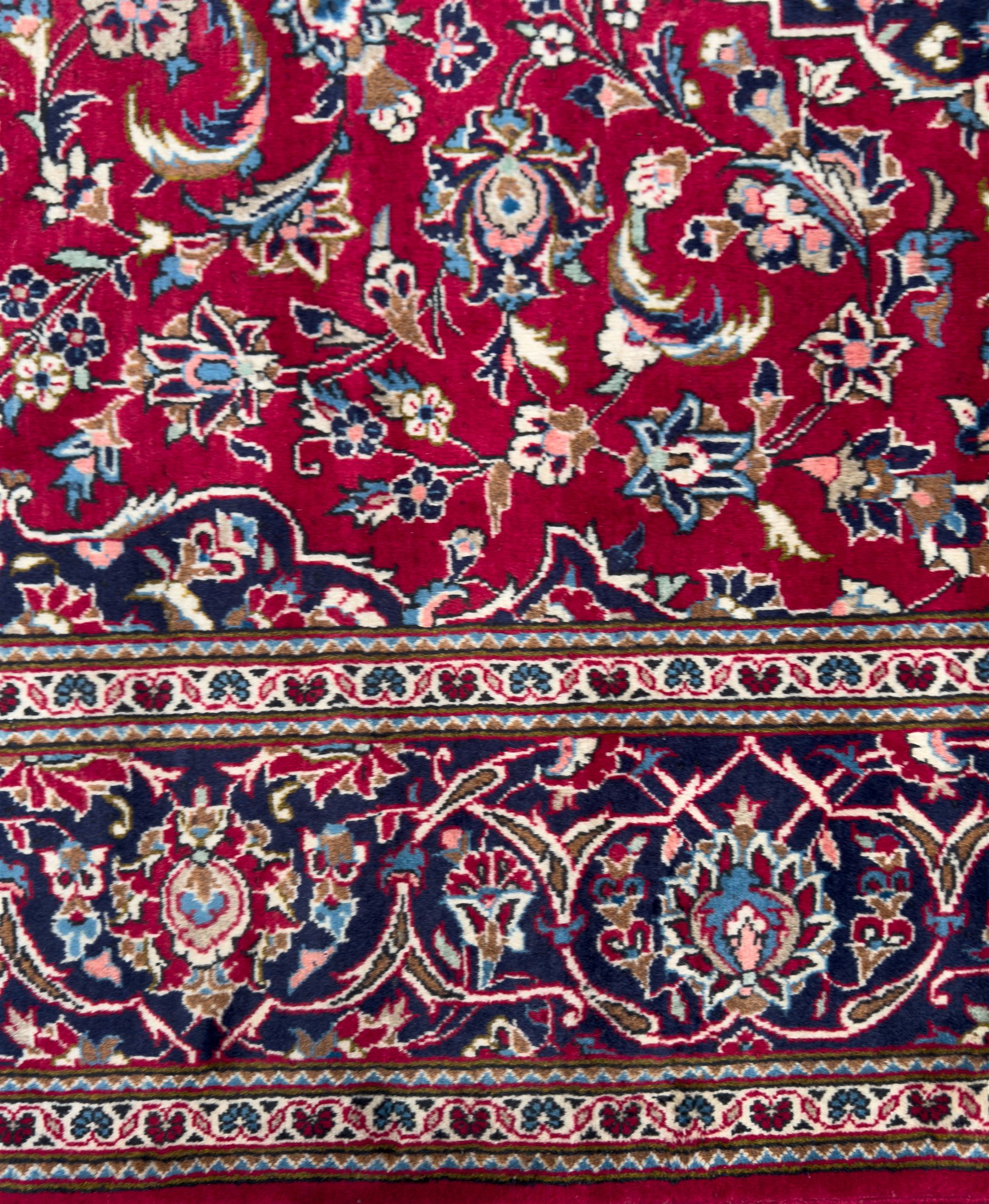 Central Persian Kashan hand-knotted carpet, the deep red ground with a central lozenge medallion in ivory and navy, surrounded by dense scrolling floral and vine motifs, navy spandrels and a wide border with stylised palmettes and flowerheads enclosed by multiple guard stripes