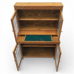 Early 20th-century Arts and Crafts glazed oak bookcase, upper section fitted with a pair of leaded glazed doors enclosing a fixed shelf above a projecting frieze drawer interior with baize-lined writing surface flanked by two compartments, carved with interlaced decoration, lower section fitted with a further pair of leaded glazed doors enclosing a single shelf, raised on short stile feet 