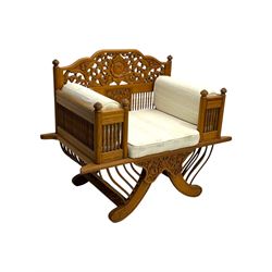 Early 20th century carved teak Howdah elephant chair, with pierced floral and foliate carved backrest, spindle gallery arm supports, cream upholstered cushions, splayed legs on a curved reeded base 