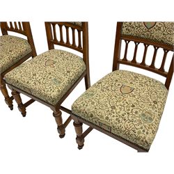 Set six Edwardian oak dining chairs, the cresting rails relief carved with foliate scrolls and pierced with handle, upholstered in foliate pattern fabric decorated with heraldic shields,. turned front supports on castors joined by plain H-framed stretchers