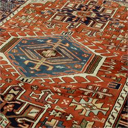 North West Persian Karajeh hand-knotted rug, terracotta ground, with a stepped geometric pole medallion design in blue, ivory and dark navy tones, surrounded by angular tribal motifs incorporating stylised flowerheads, hooked devices and geometric vine work, enclosed by a dark navy border with repeating rosettes and angular leaf motifs, flanked by narrow patterned guard stripes, with cotton fringes